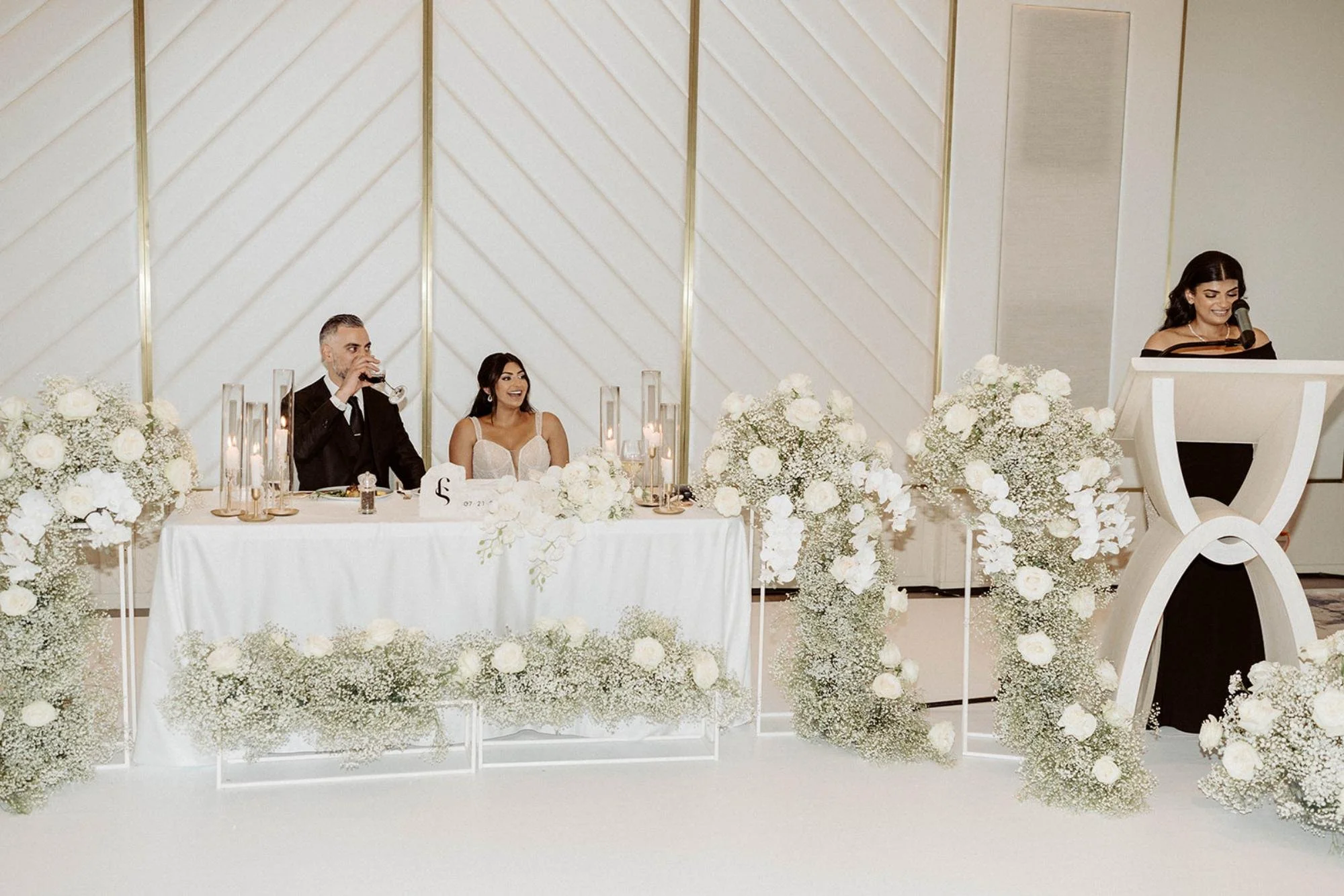 Bride and groom seated at their sweetheart table decorated with white flowers, listening to a speech from a woman standing at a podium.