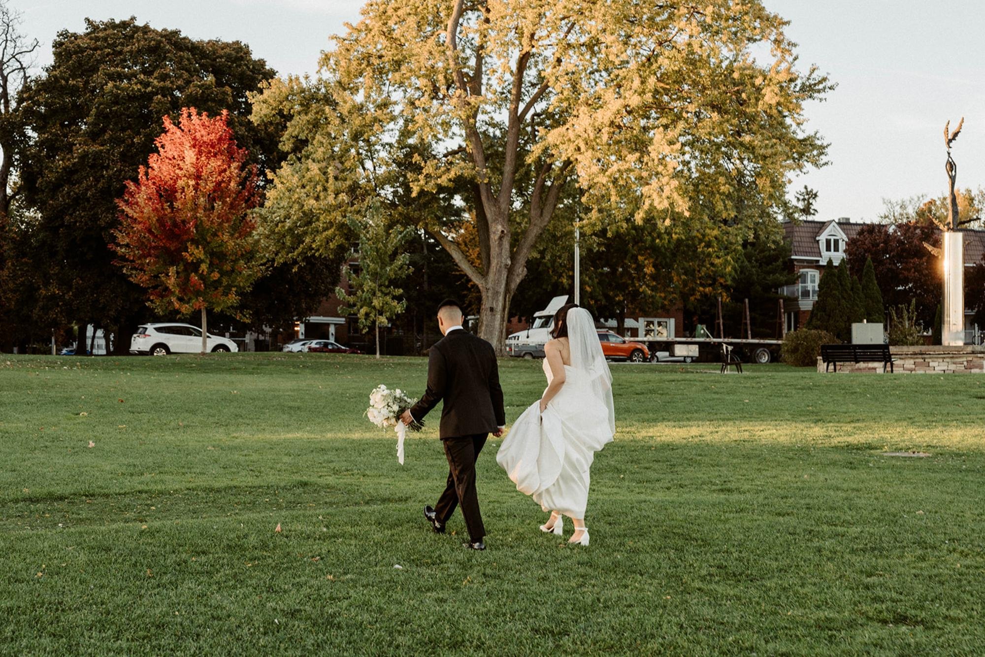 Bride and groom walk across a grassy park surrounded by colorful autumn trees.