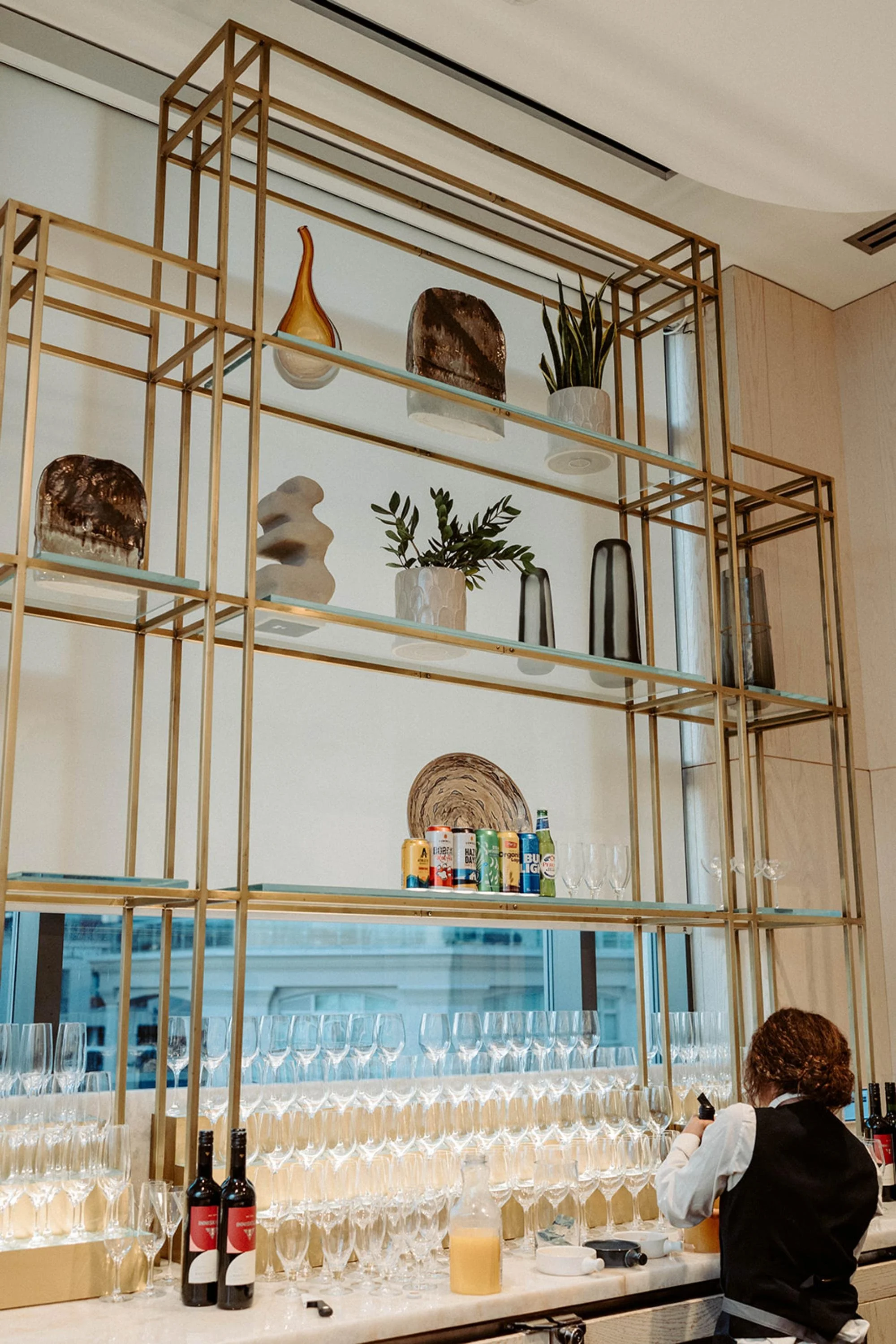 Back bar display with gold shelving, decorative pieces, wine glasses, and a bartender preparing drinks.