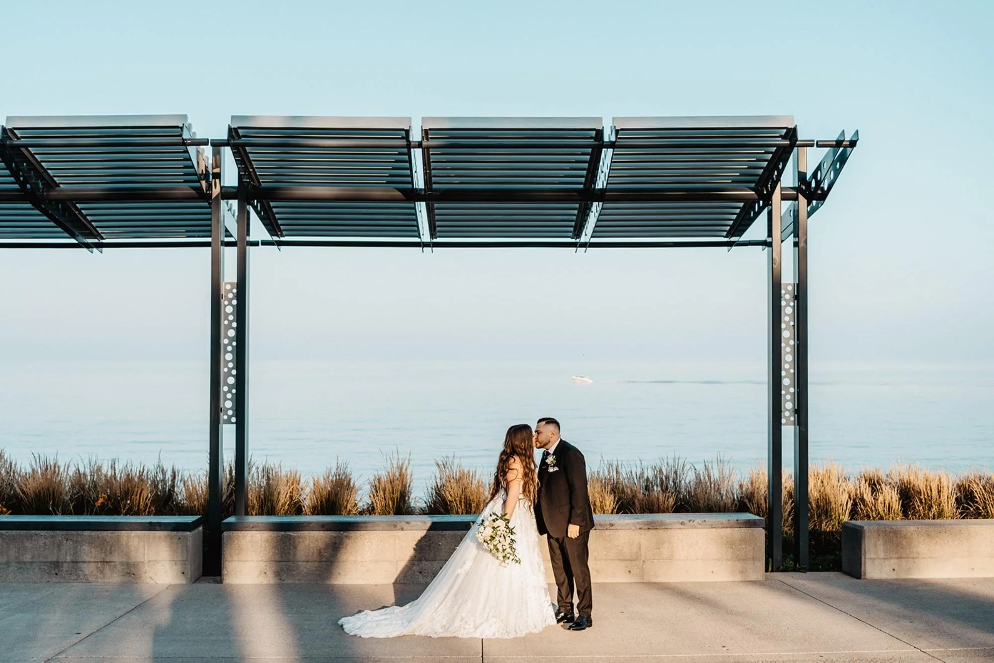 Bride and groom share a kiss under a metal pergola overlooking the calm waterfront.