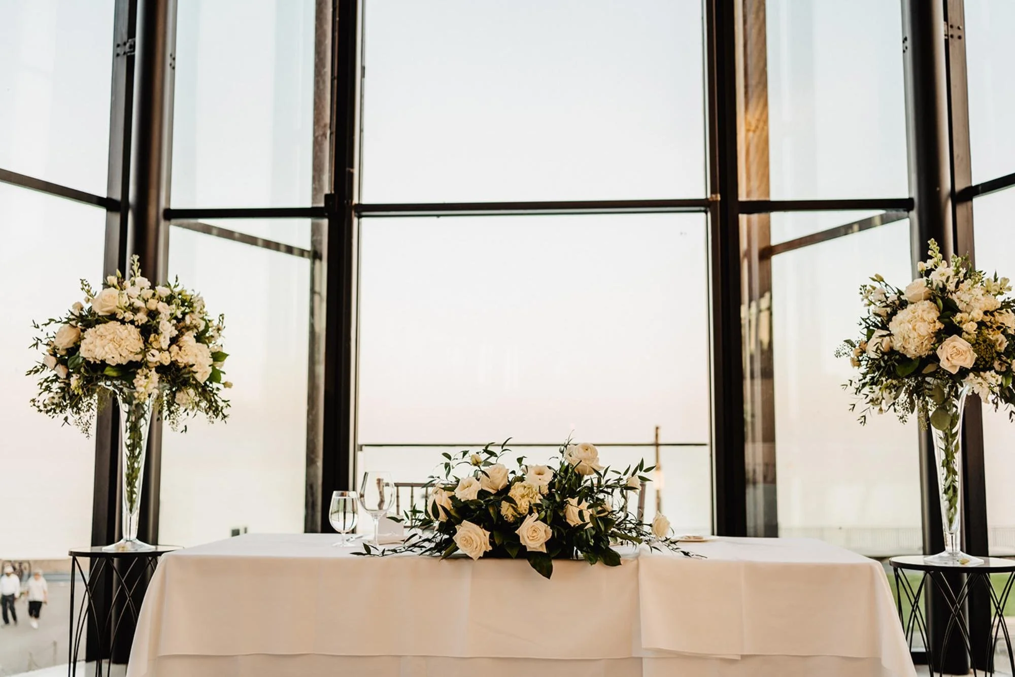 Sweetheart table decorated with white floral arrangements in front of large windows.