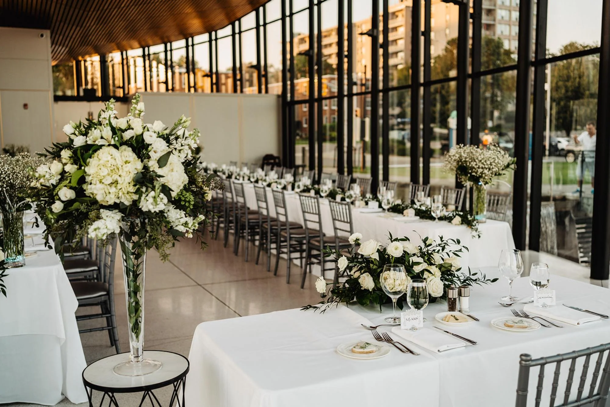 Reception hall with long tables, white linens, and tall floral centerpieces near floor-to-ceiling windows.