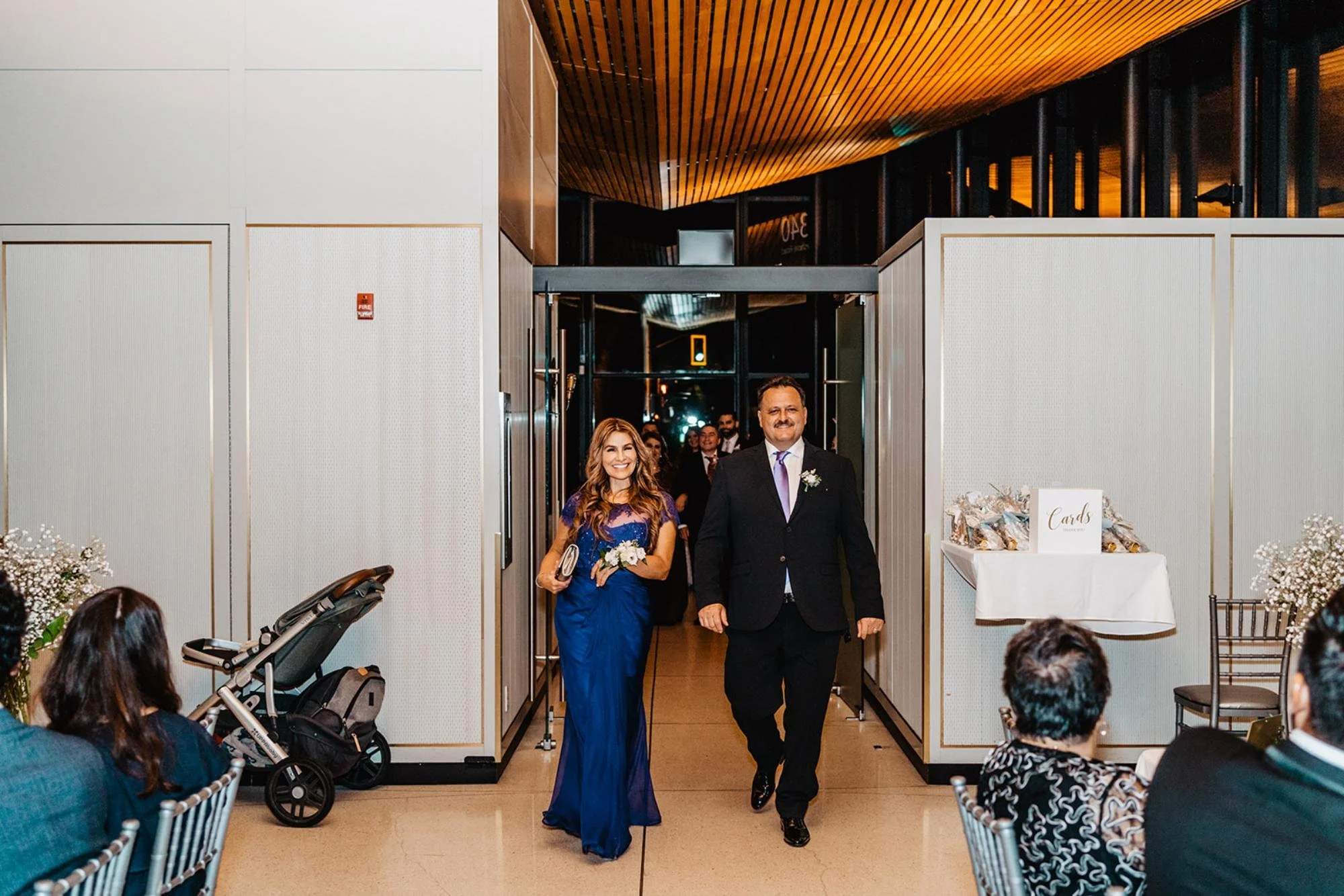 Two formally dressed guests walk into the reception as others look on and smile.