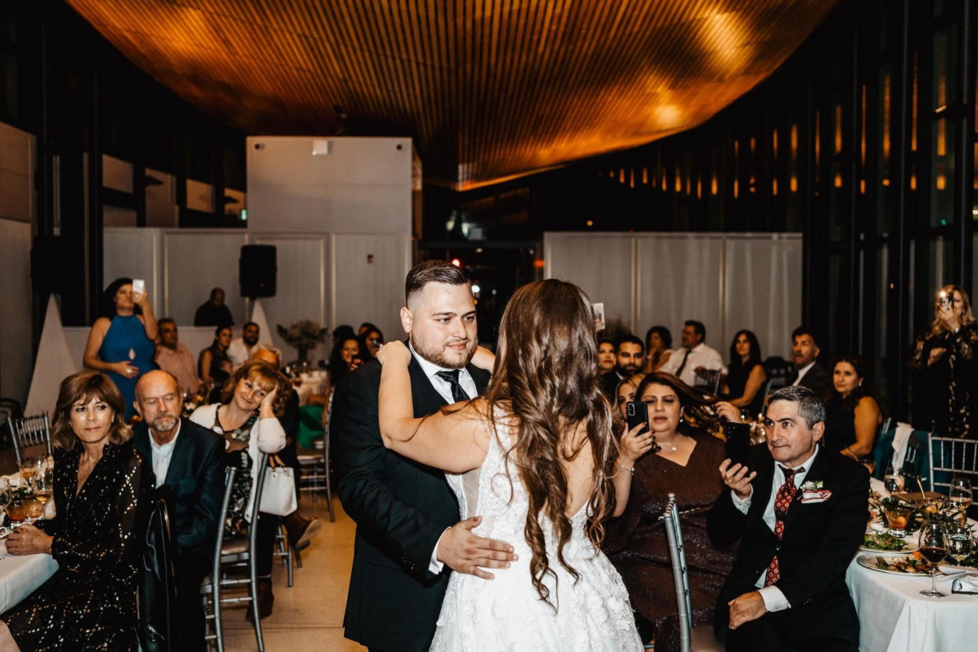 Bride and groom share their first dance while guests watch and record the moment.