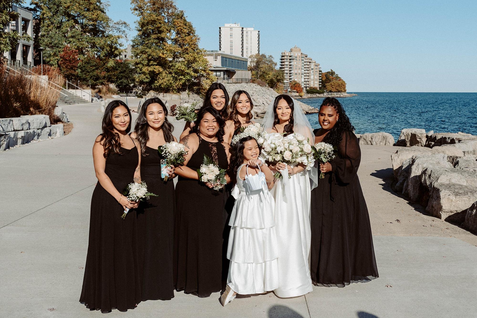 Bride and bridesmaids in black dresses pose by the waterfront on a sunny day.