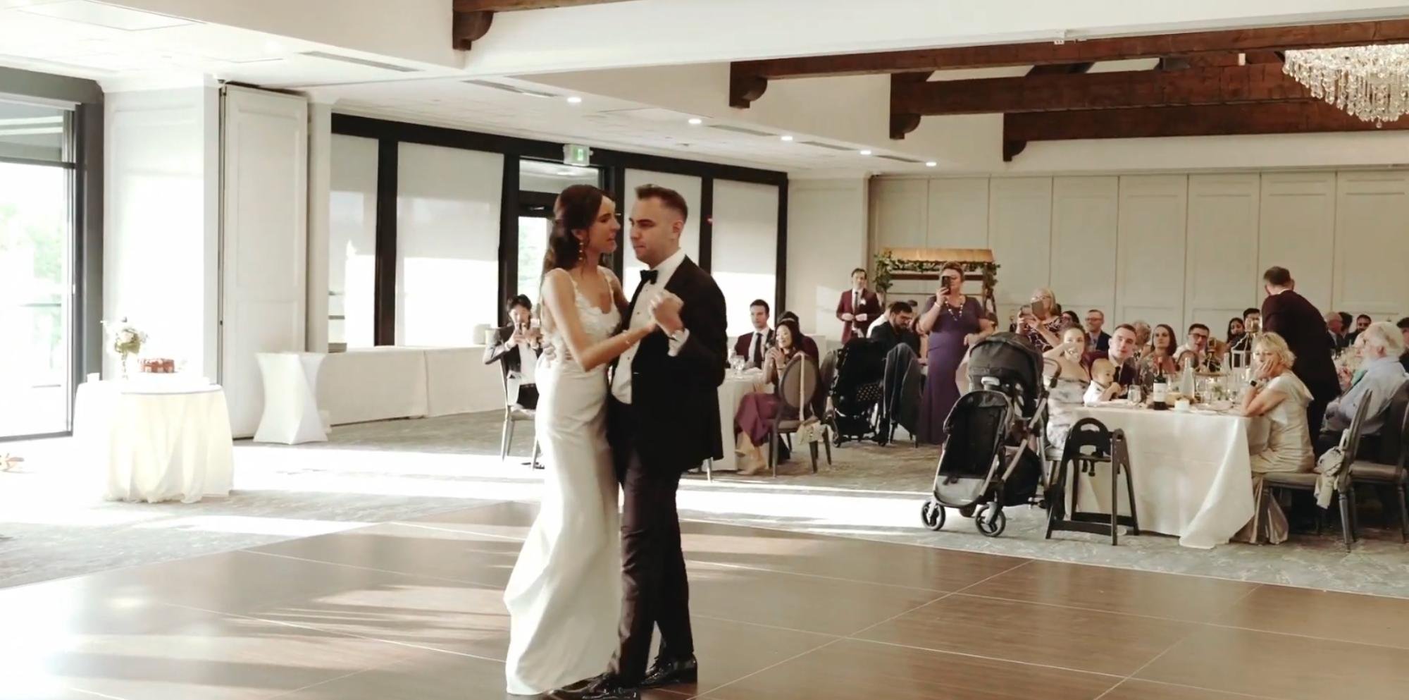 The bride and groom share their first dance on a wooden floor in a sunlit reception hall while nearby guests watch, clap, and record the moment.