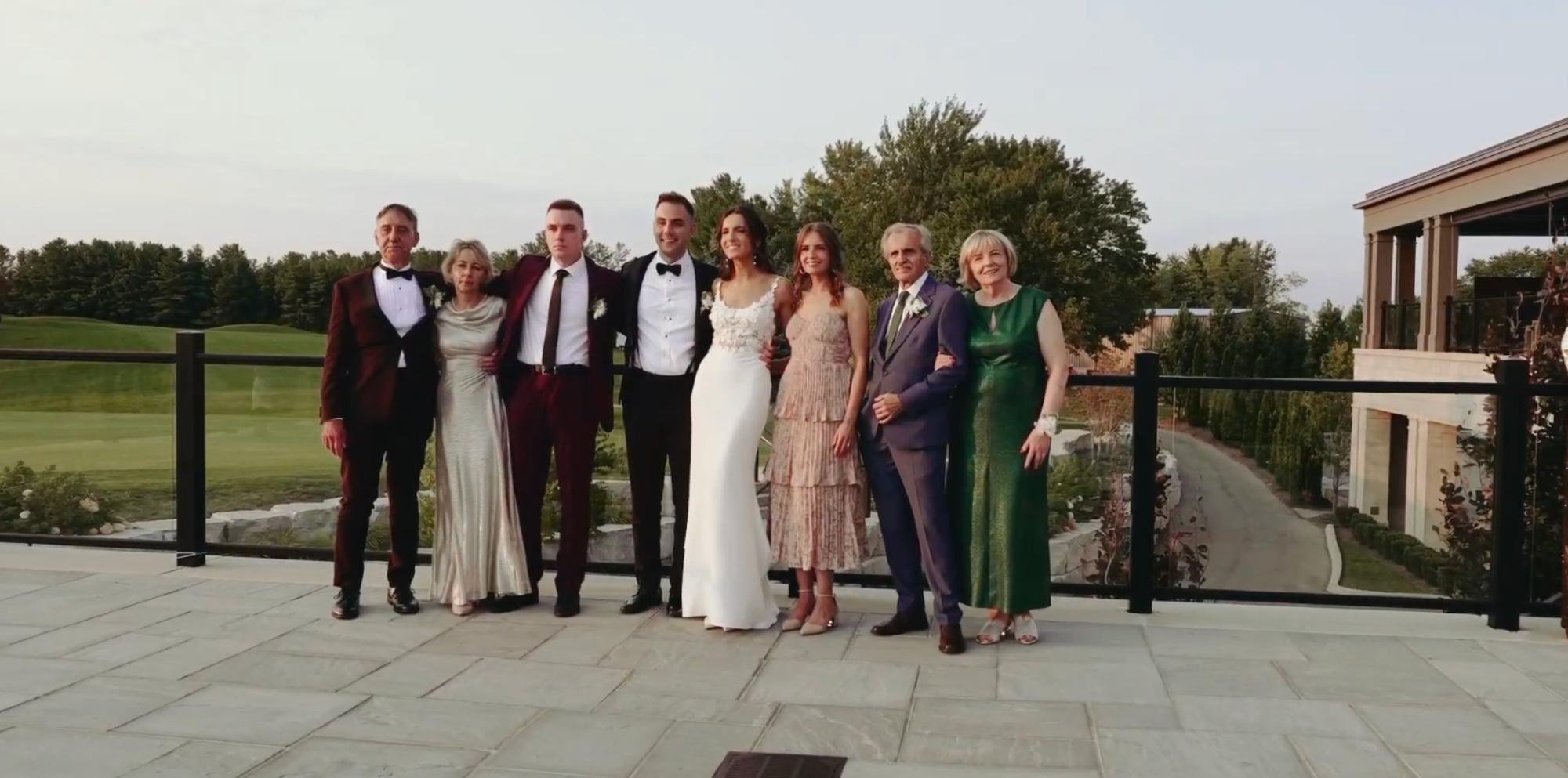 A large wedding party poses outdoors on a terrace at sunset, dressed in suits and elegant dresses, with a golf course and trees behind them.