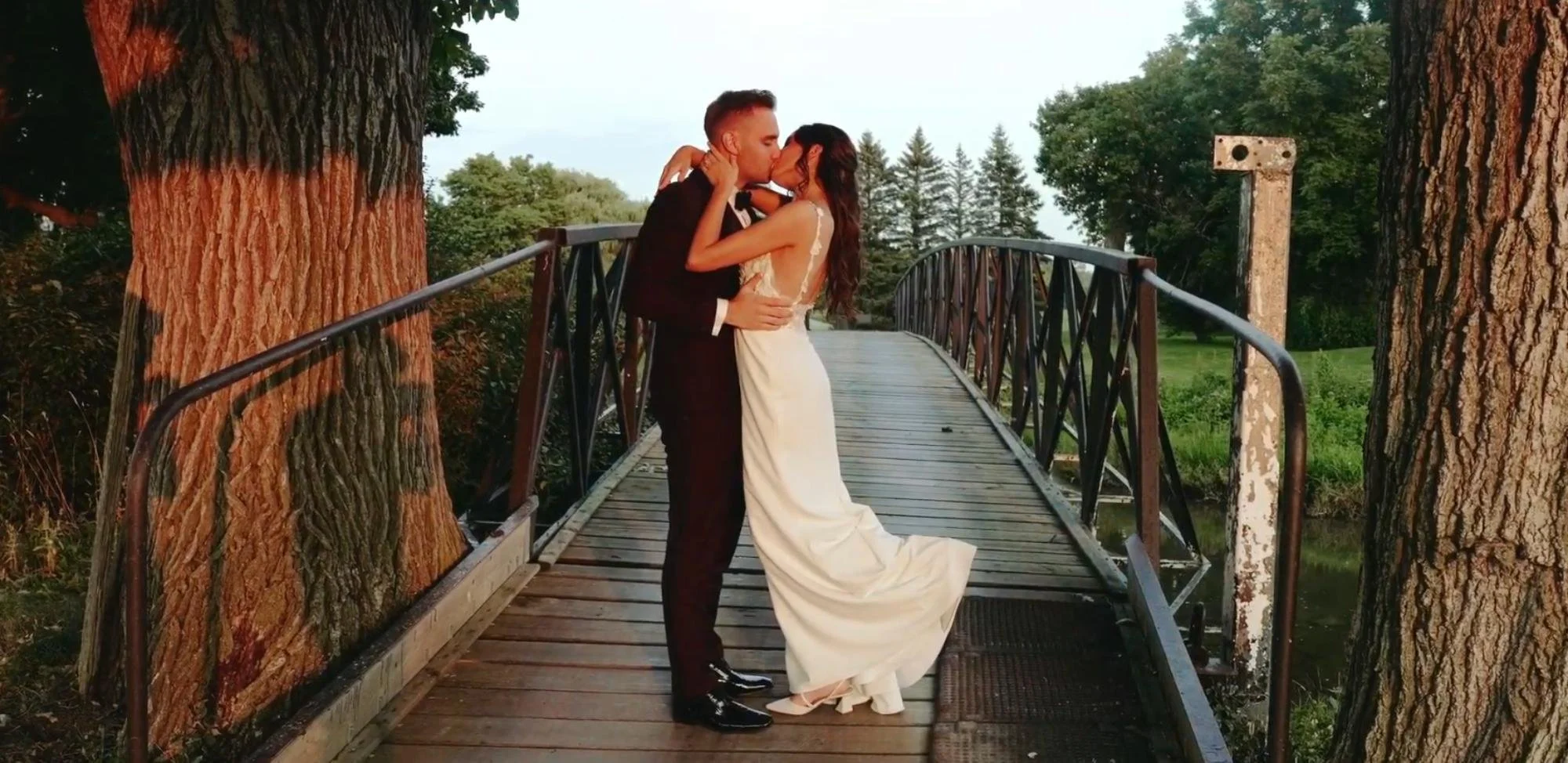 The newlyweds kiss on a wooden bridge beside a large tree, the bride’s dress flowing behind her.