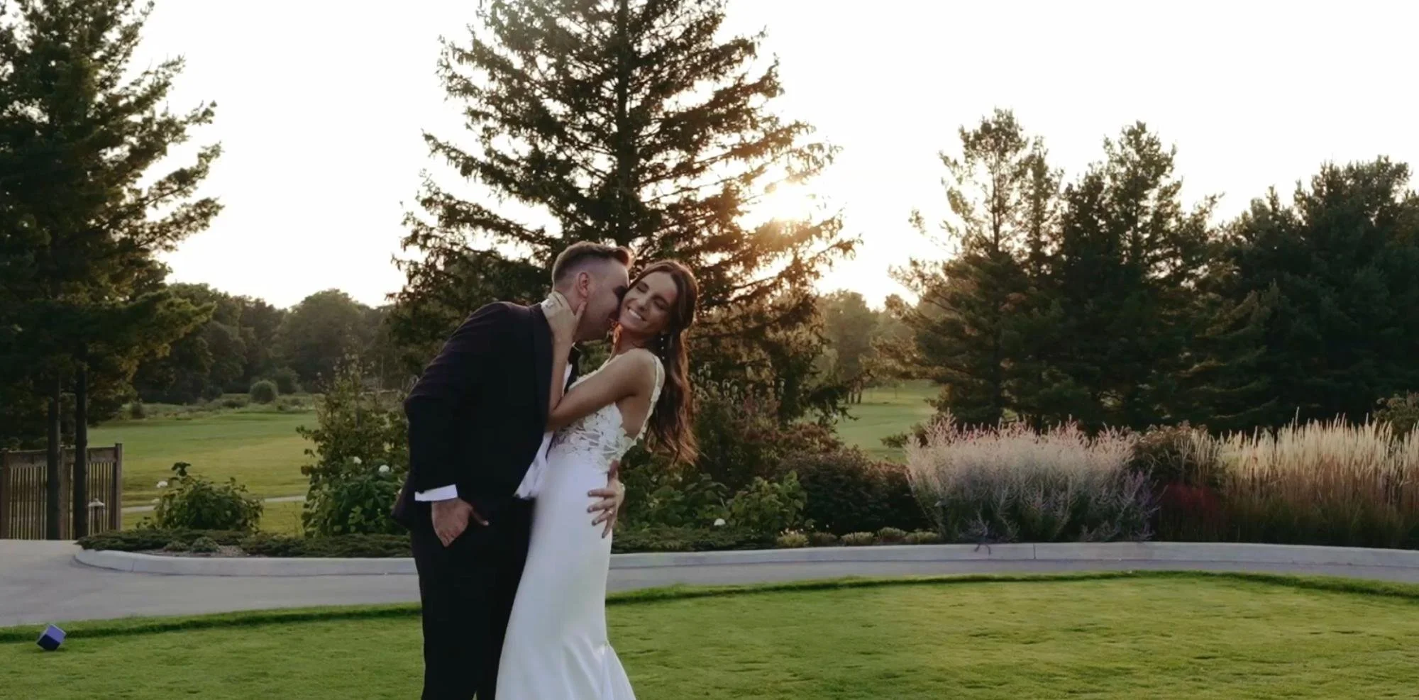 The bride and groom laugh and hold each other on a grassy area at sunset, with tall trees and ornamental plants in the background.