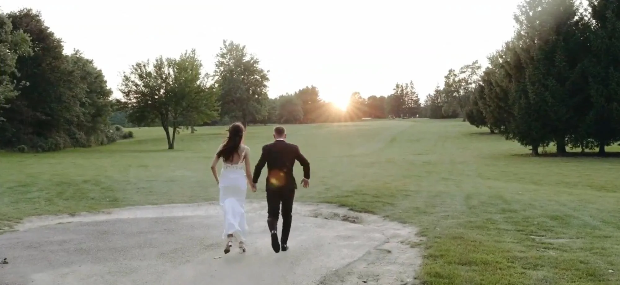 The couple runs hand-in-hand across an open grassy field toward the setting sun, framed by trees on both sides.