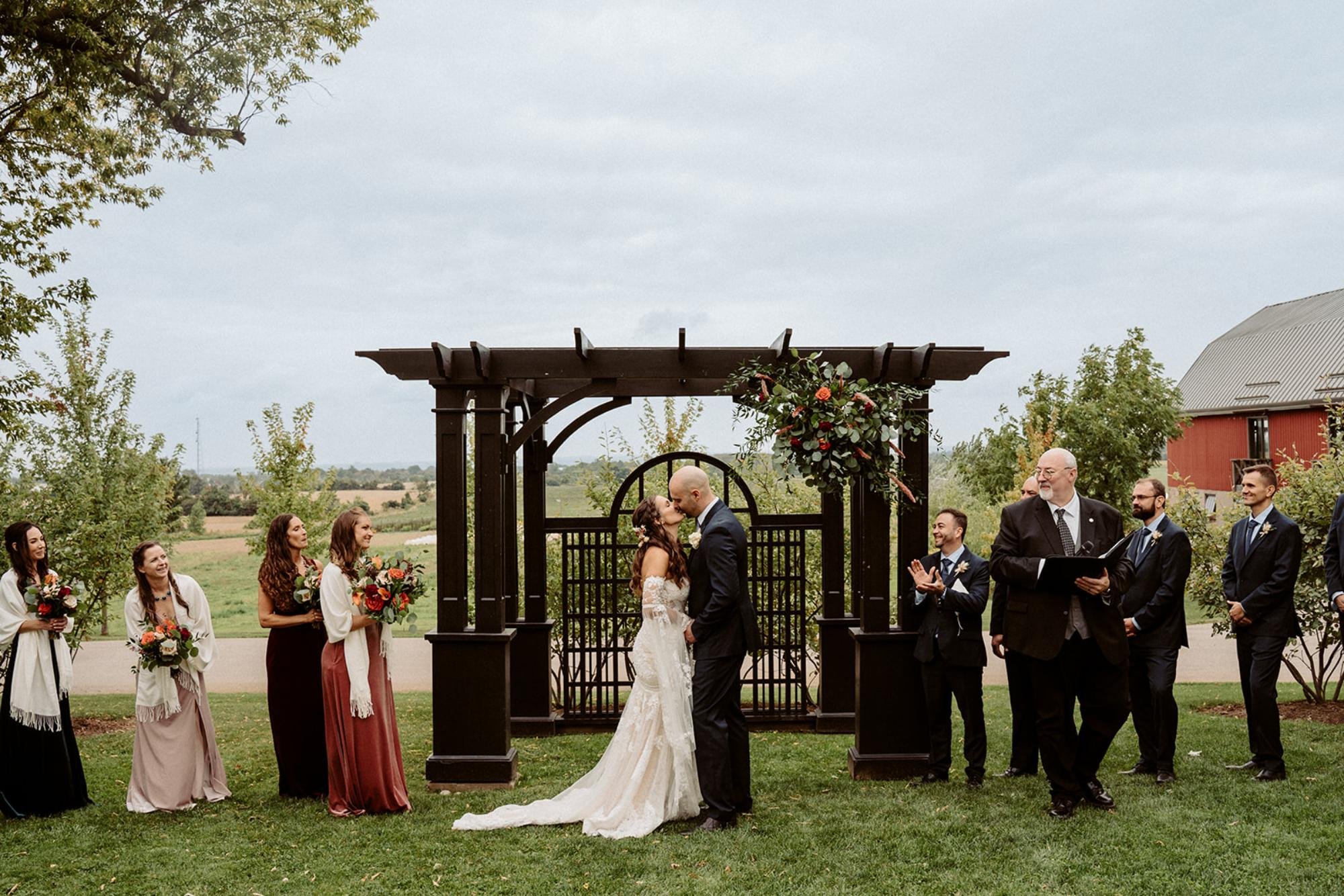 Bride and groom kiss under a floral-decorated pergola during the outdoor ceremony.
