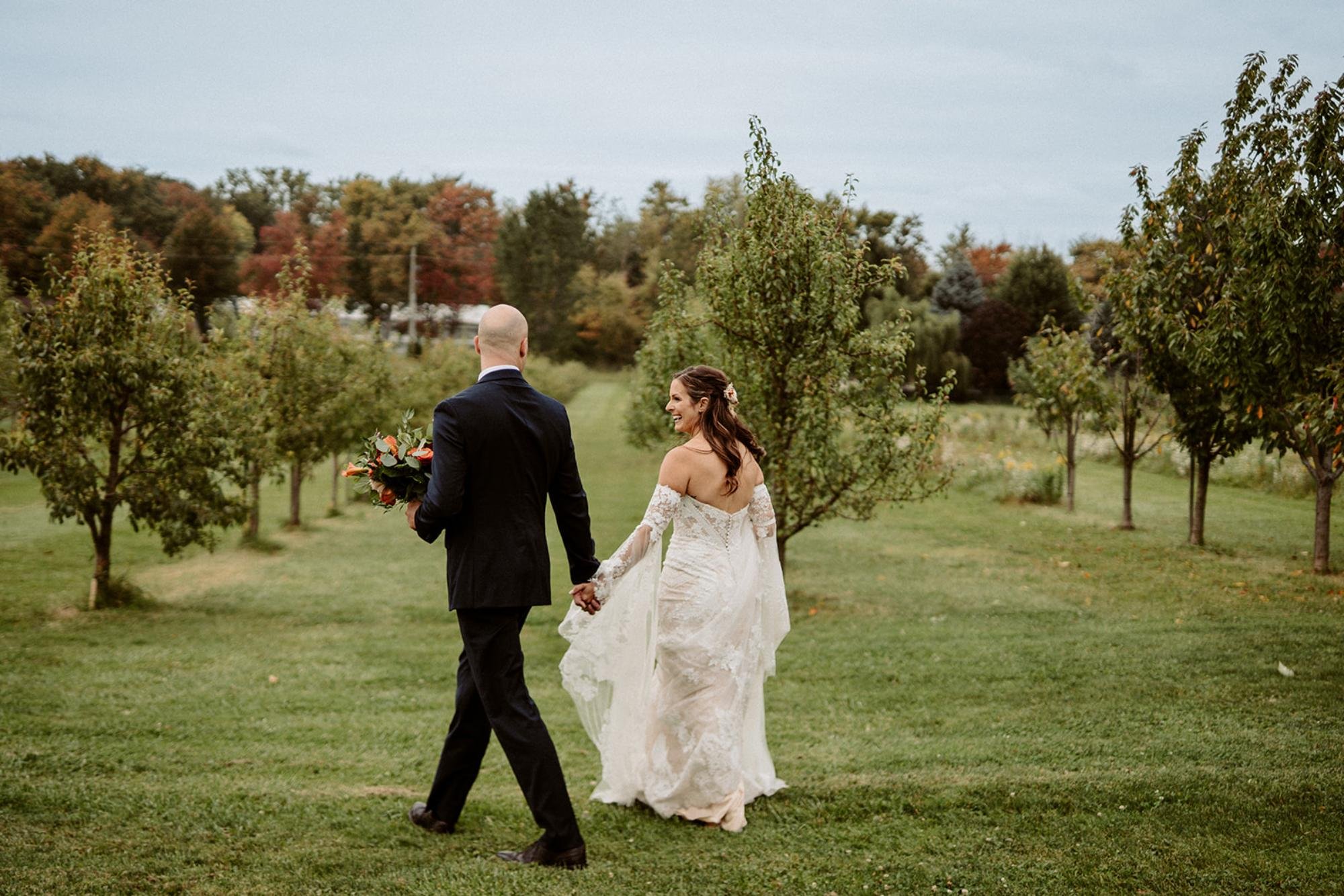 Bride and groom walk hand-in-hand through an orchard with fall trees in the background.