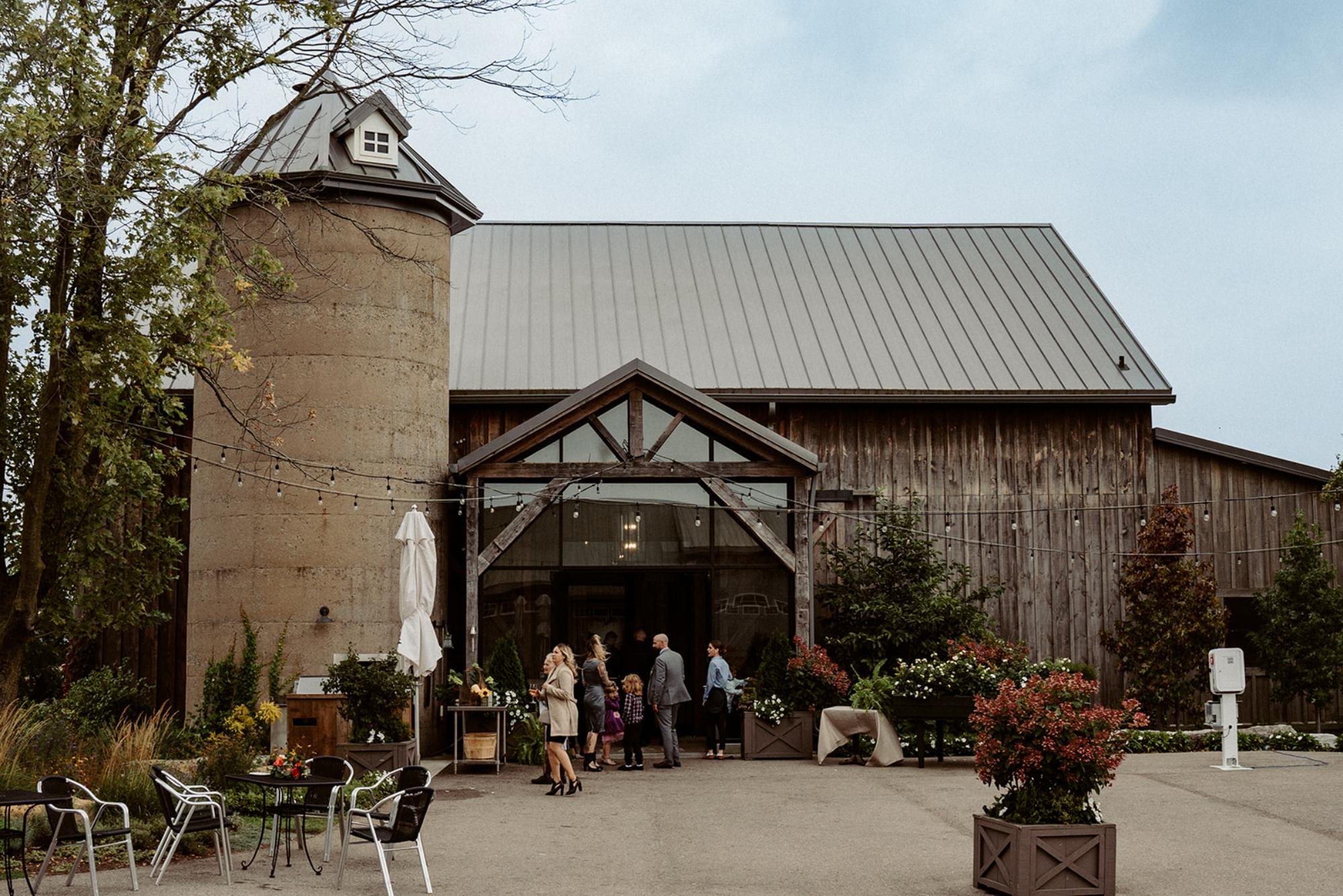 Guests gather at the entrance of a rustic barn with a silo and string lights.