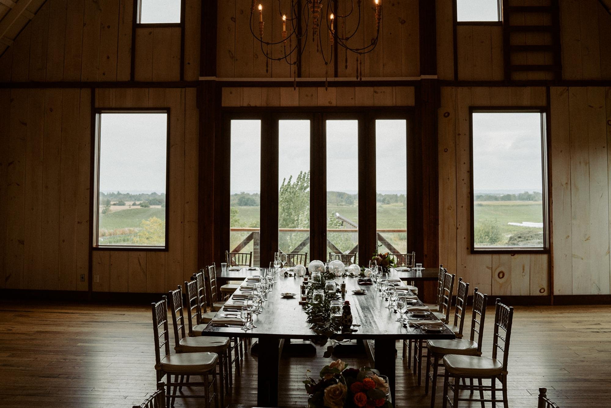 Reception head table set inside the barn, framed by tall windows overlooking the fields.