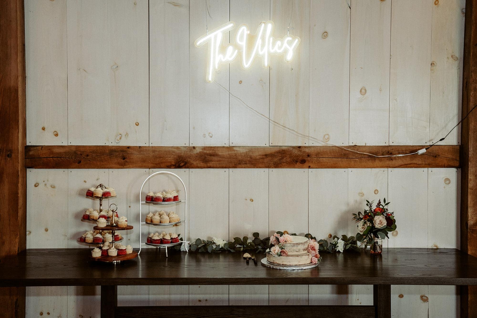 Dessert table with cupcakes, a floral cake, and a neon sign reading “The Miles.”