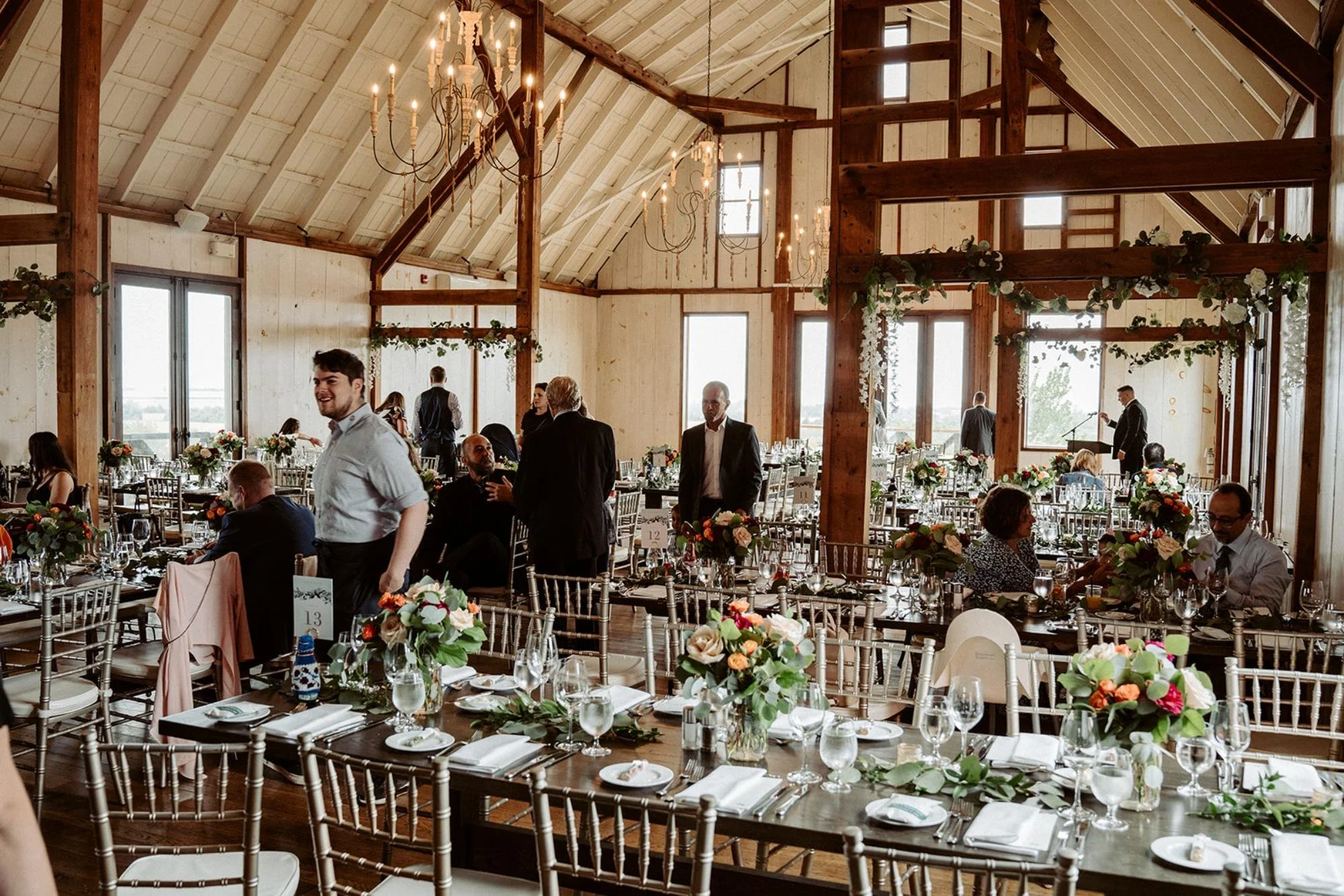 Guests mingle and find their seats in a decorated barn reception hall with chandeliers.