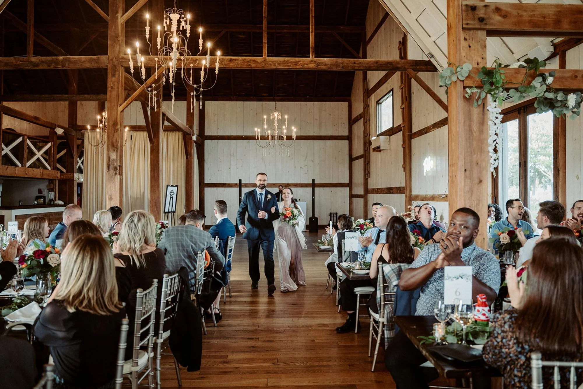 Wedding party members enter the reception as guests applaud from both sides.