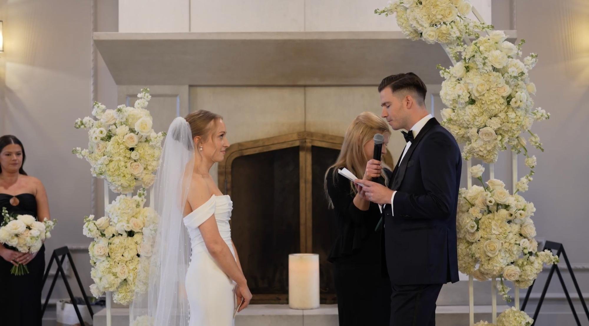A bride and groom exchange vows indoors, standing between tall white floral arrangements.