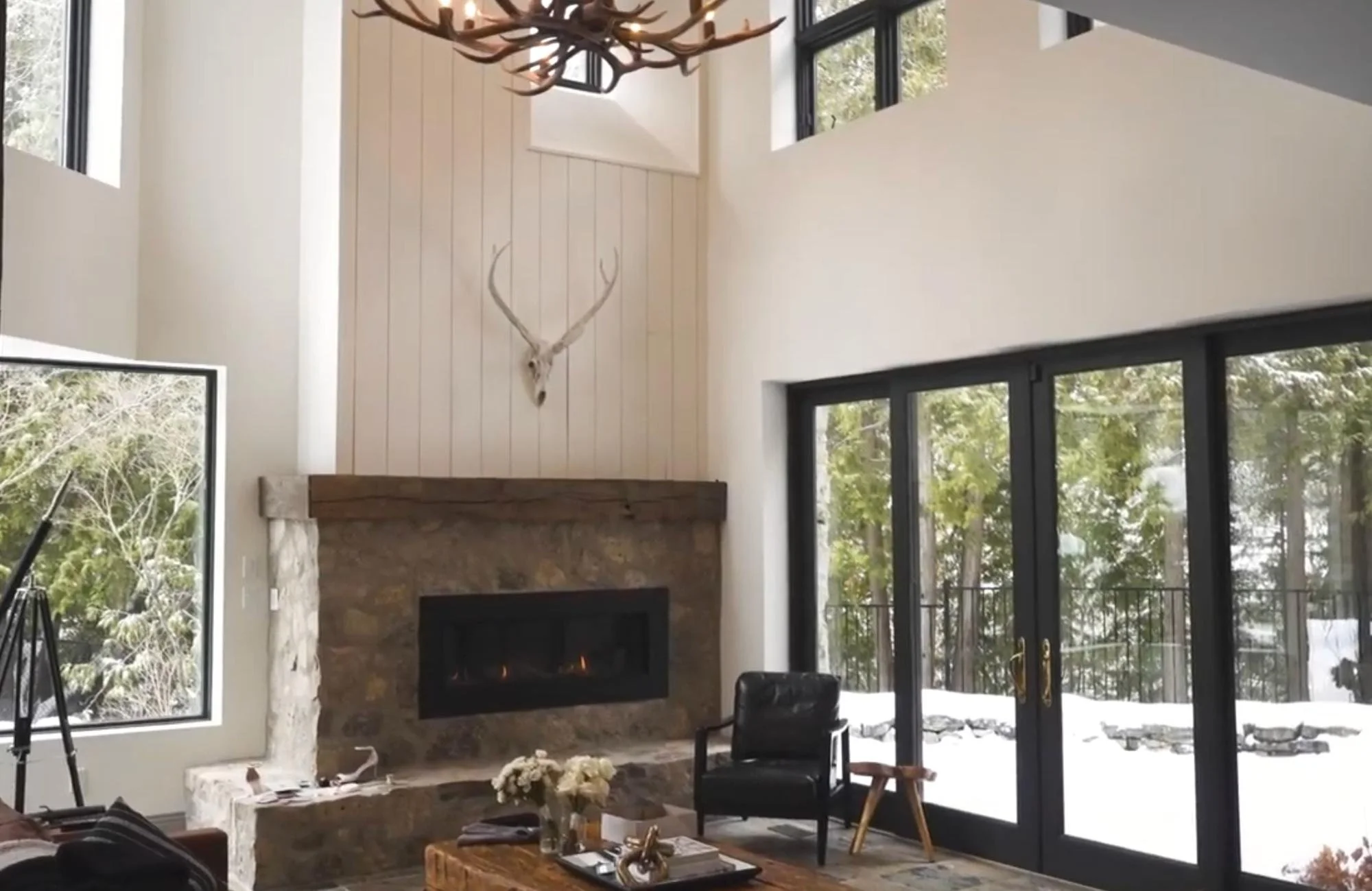 A cozy living room with a stone fireplace, antler chandelier, and snowy forest views through large windows.