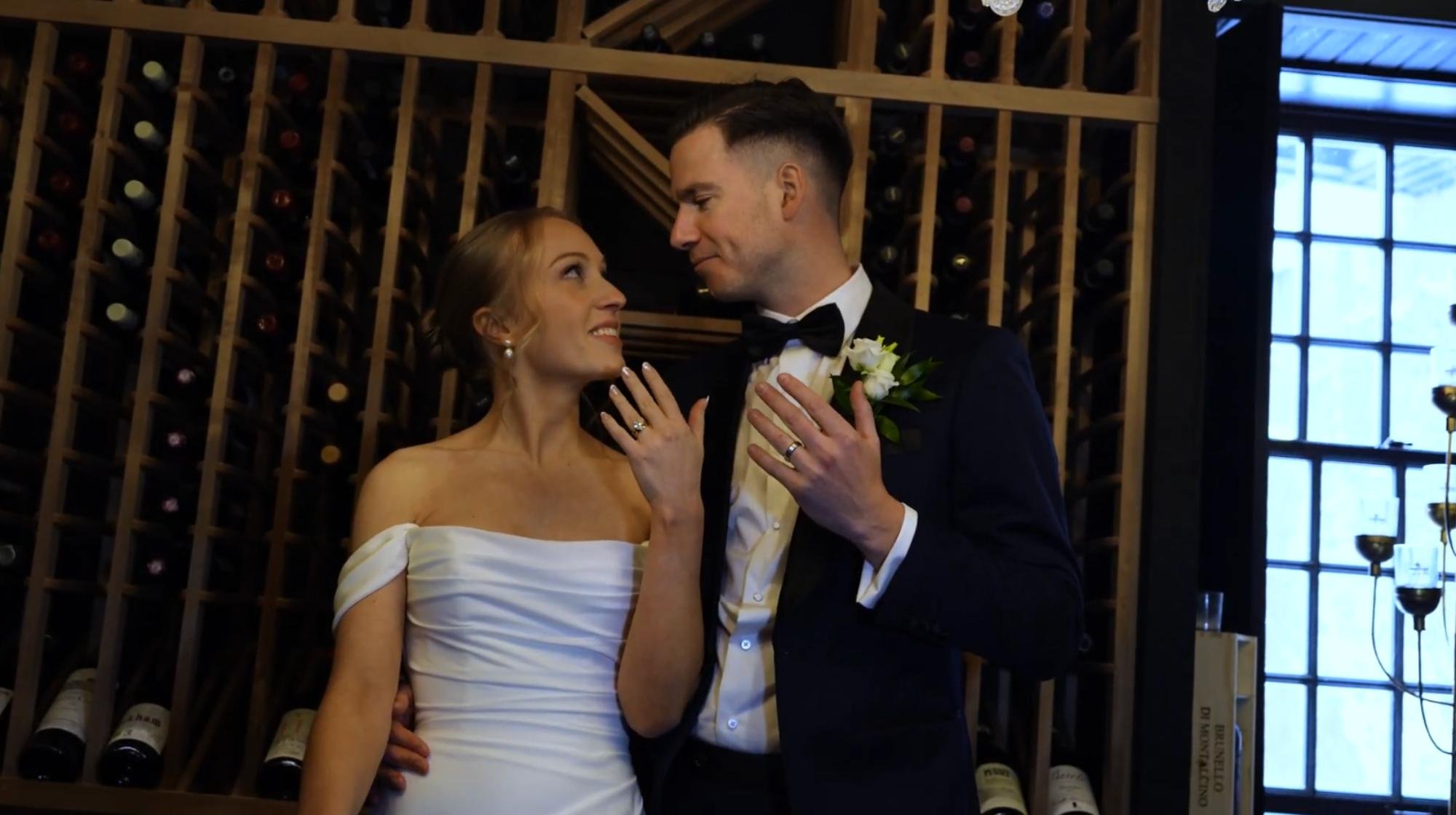 The newlyweds smile at each other and show their rings while standing in a wine cellar.