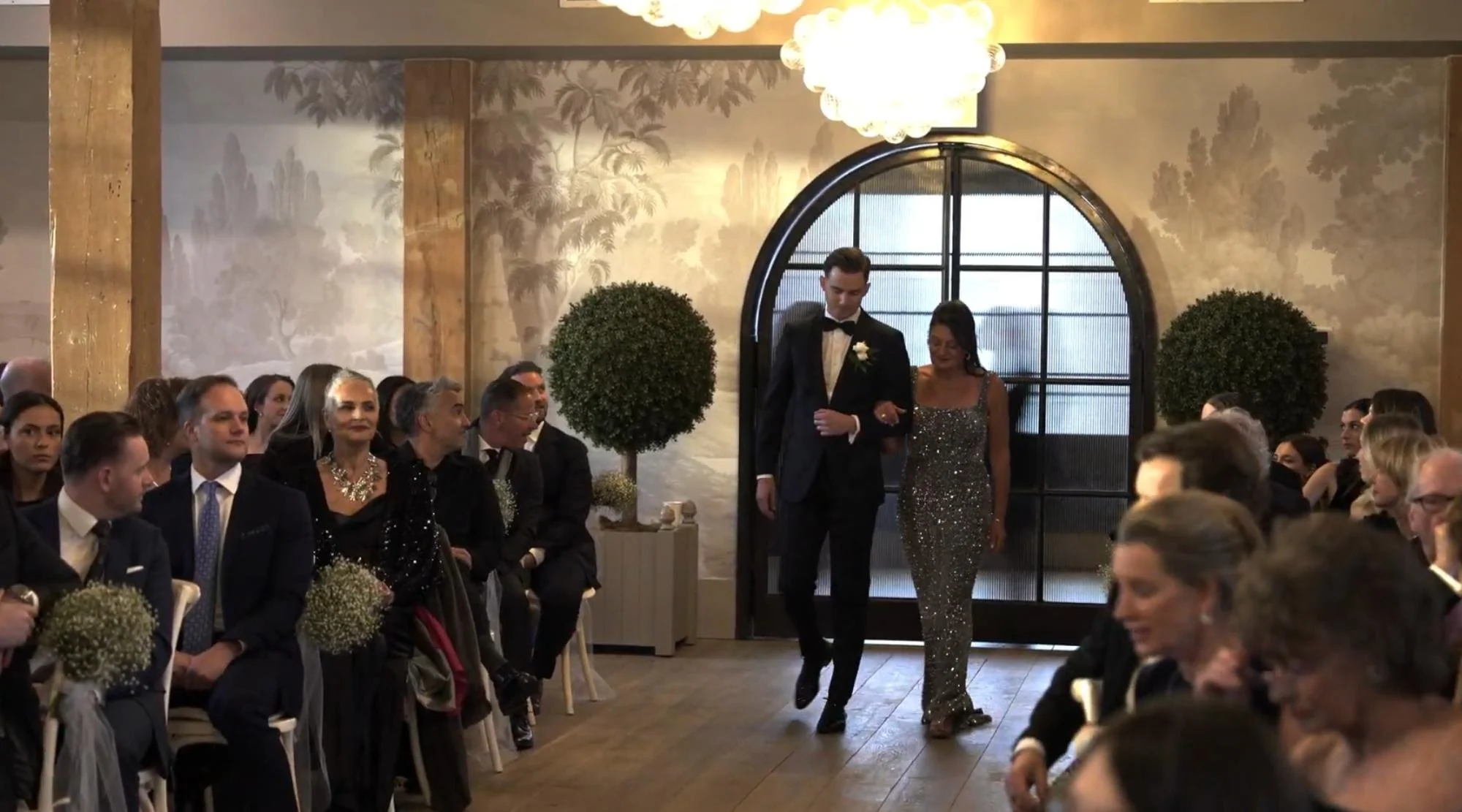 A man in a suit walks down the aisle with a woman in a sparkly gown as guests look on.