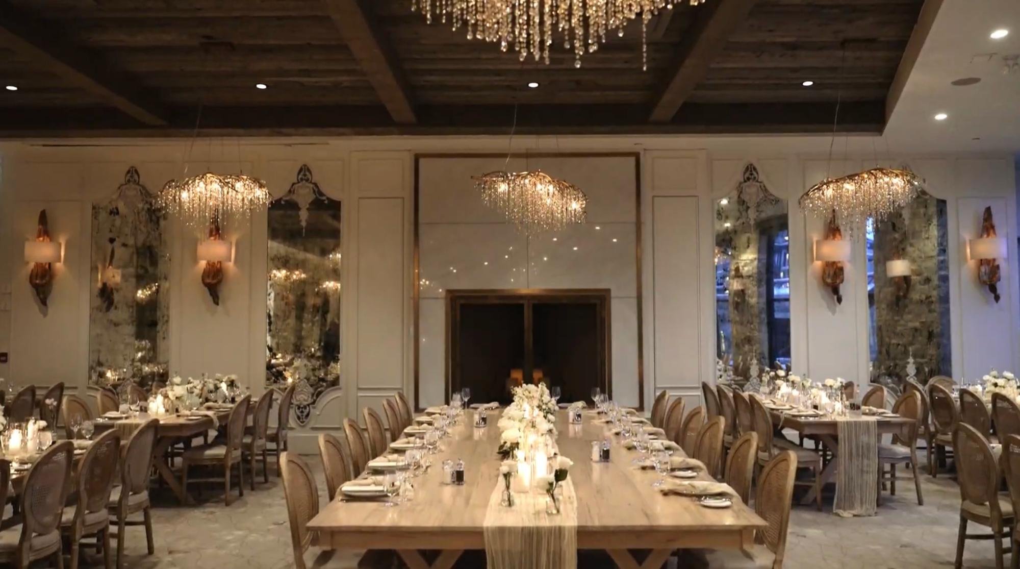 A long reception table is set with candles and white flowers beneath elegant chandeliers.