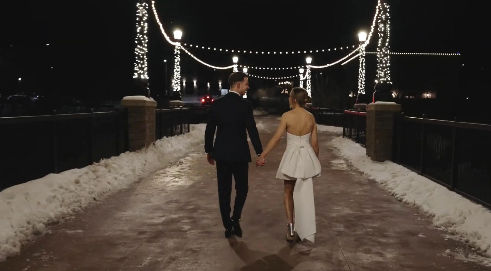 The couple walks hand-in-hand down a snowy, lit walkway at night.