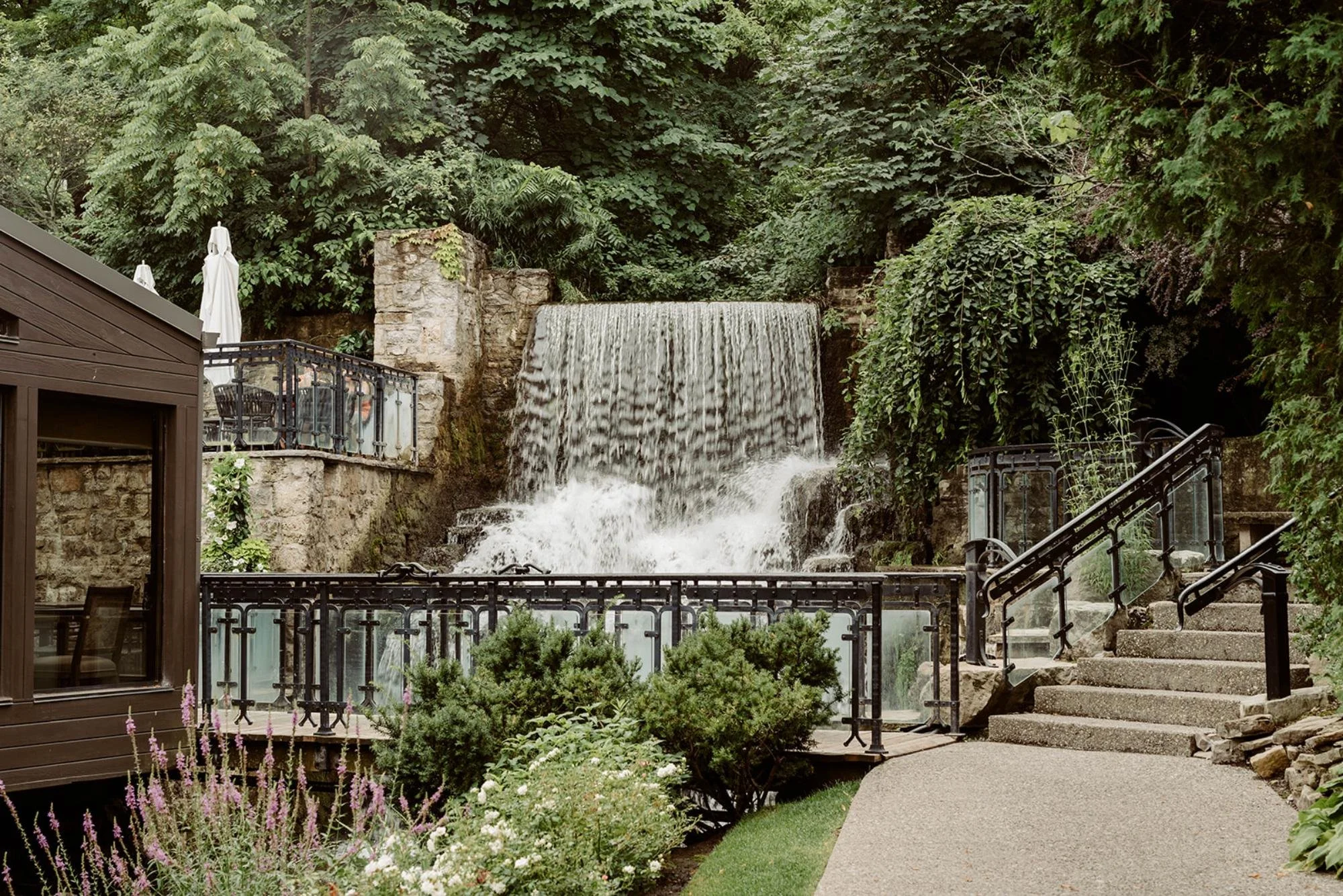 Scenic waterfall surrounded by stone walls, lush greenery, and garden pathways with metal railings.