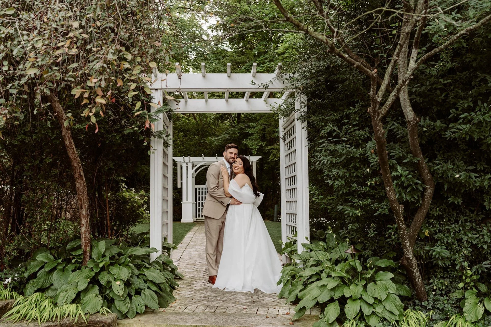 Bride and groom embracing under a white garden arbor walkway framed by lush greenery.