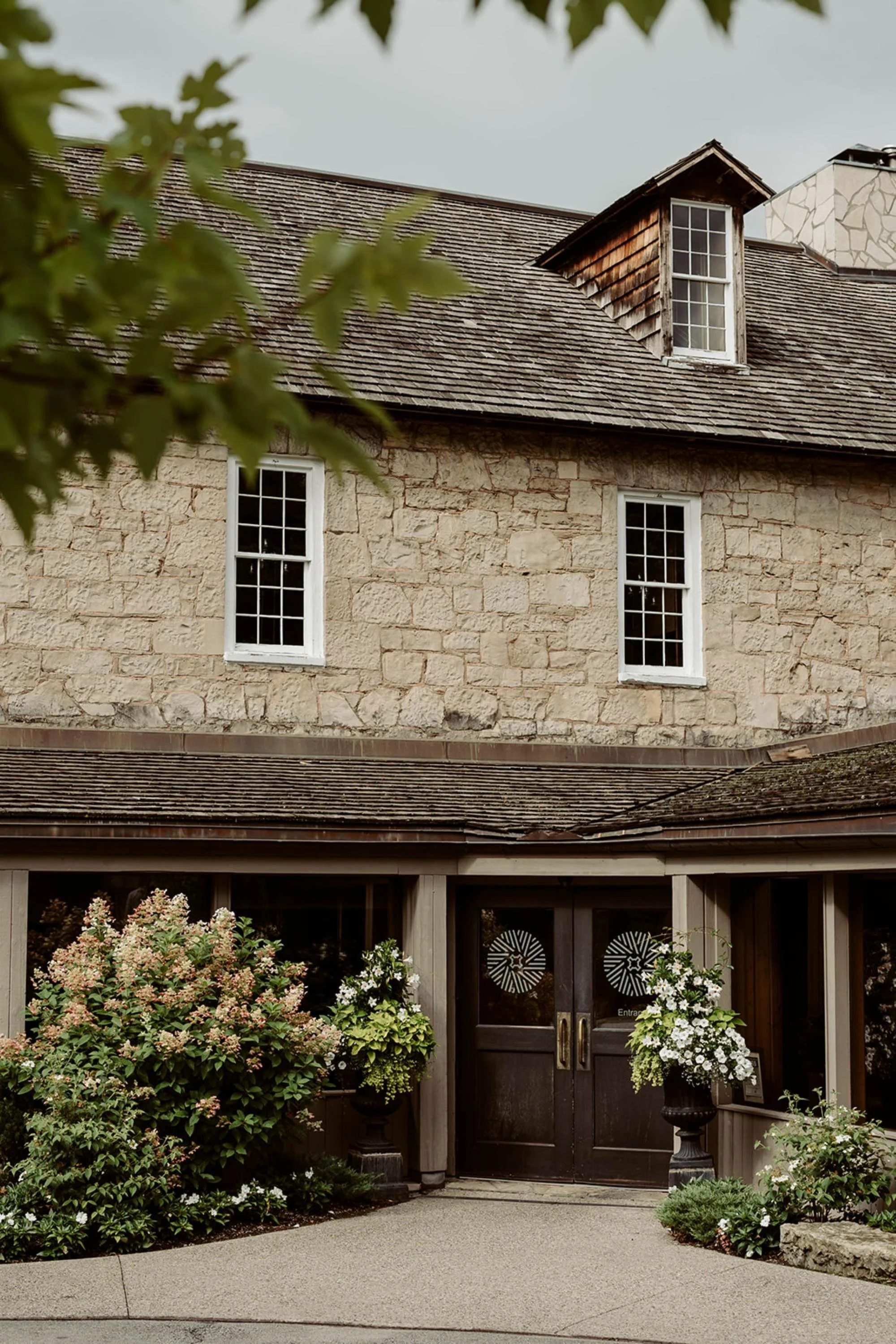 Exterior view of a historic stone building with large windows, surrounded by greenery and blooming plants.