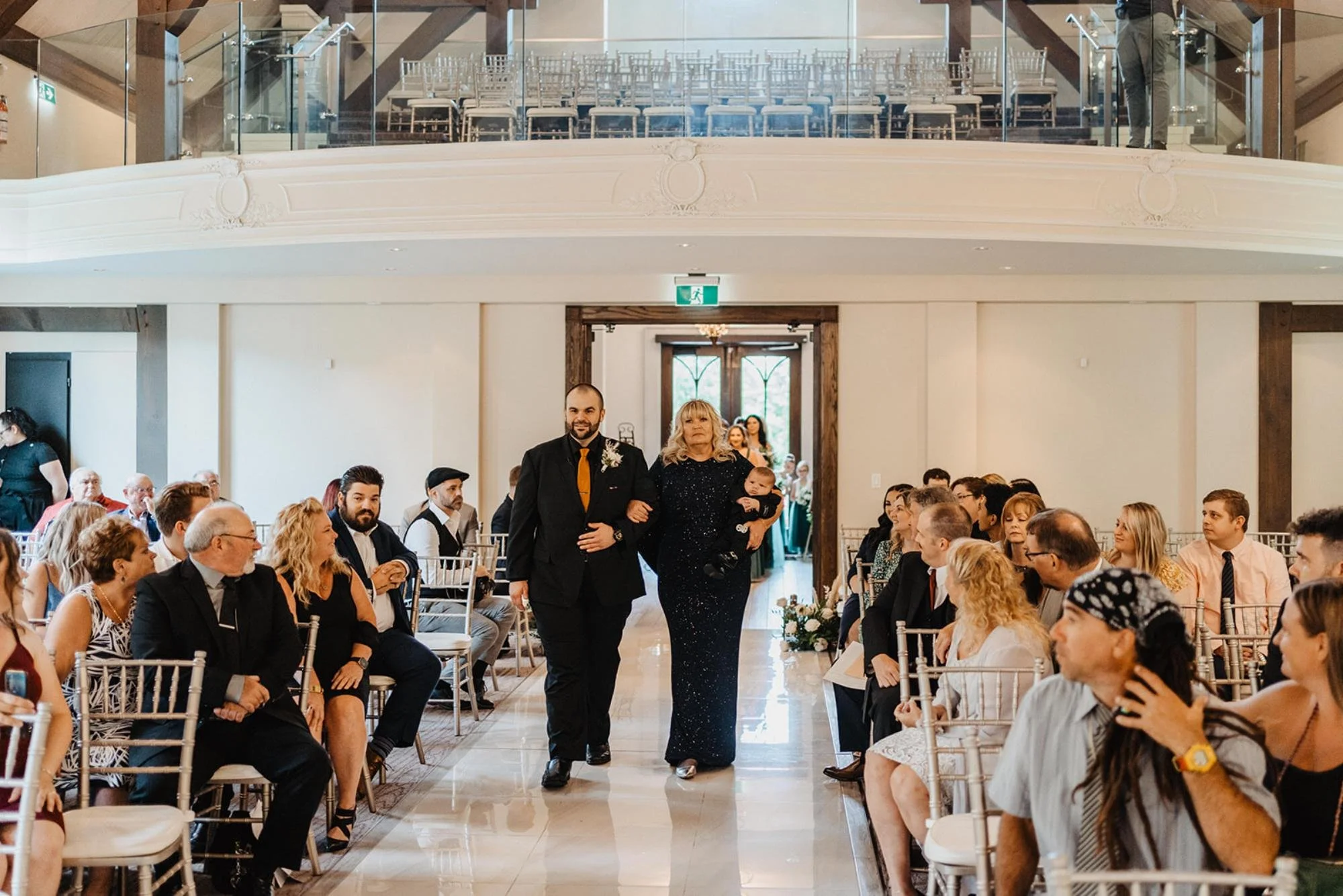 Wedding guests seated as a man escorts a woman carrying a baby down the aisle inside a bright ceremony hall with a balcony.