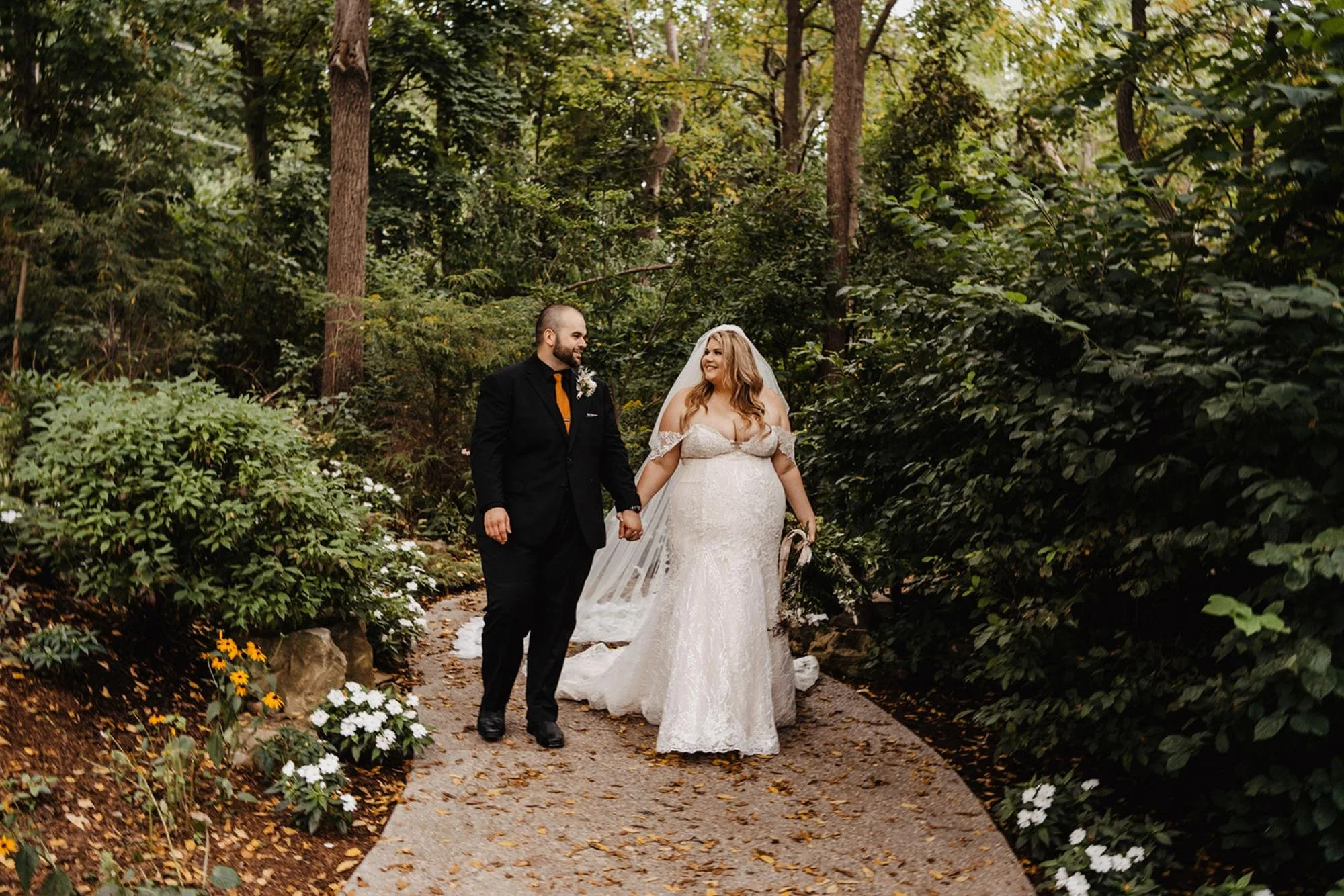 Bride and groom walking hand in hand down a garden path surrounded by trees, greenery, and flower beds.