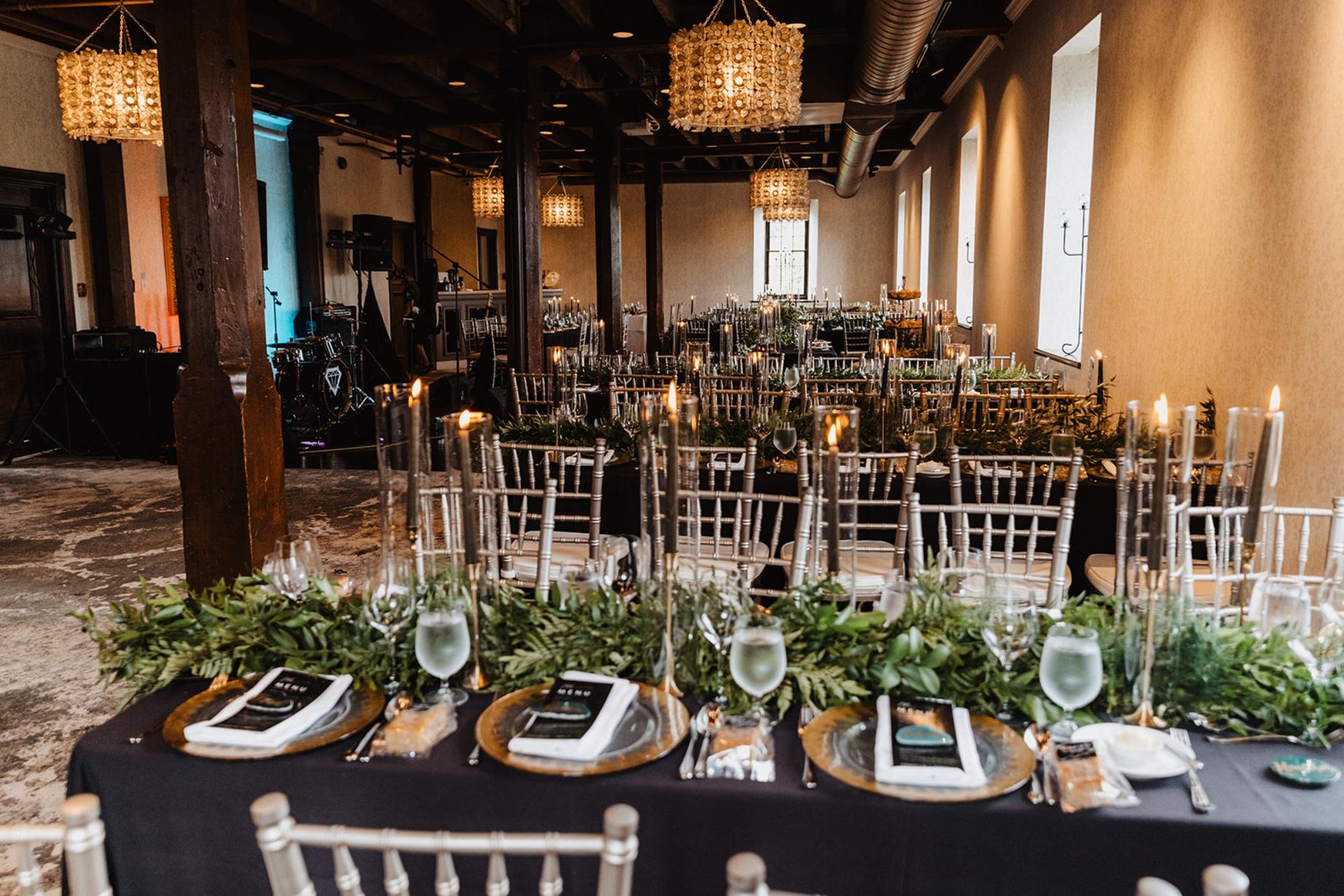 Wedding reception tables set with gold chargers, tall candles, greenery runners, and chiavari chairs in a dim, elegant banquet hall.