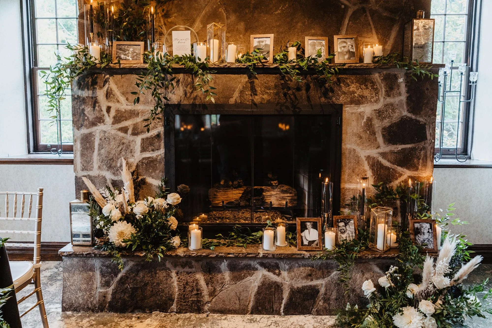 Stone fireplace decorated with candles, greenery, framed photos, and floral arrangements for a memorial or tribute display.