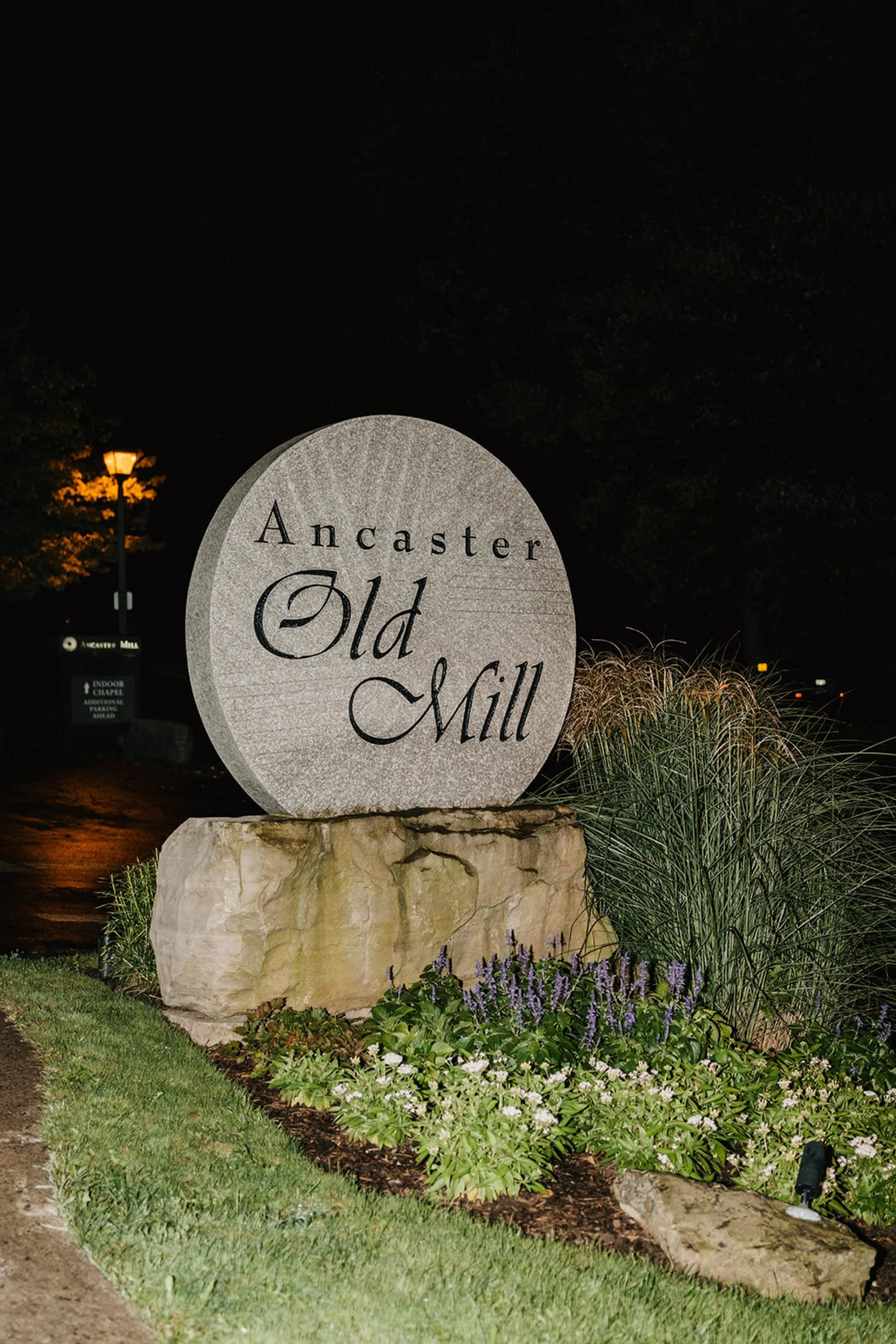 Large engraved stone sign reading “Ancaster Old Mill” surrounded by garden plants, lit at night.