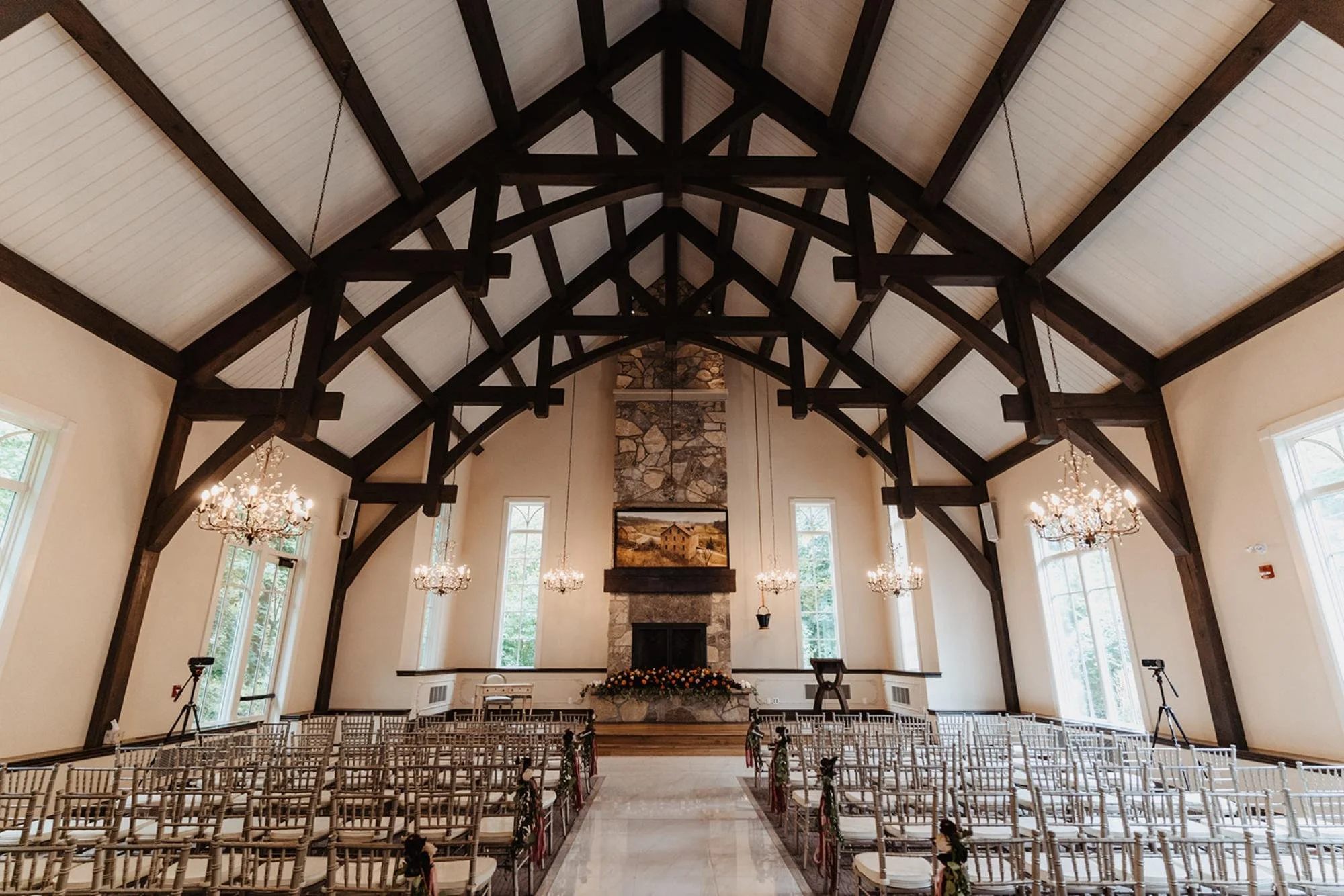 Indoor ceremony space with high wooden beams, chandeliers, rows of chairs, and a stone fireplace decorated with flowers.