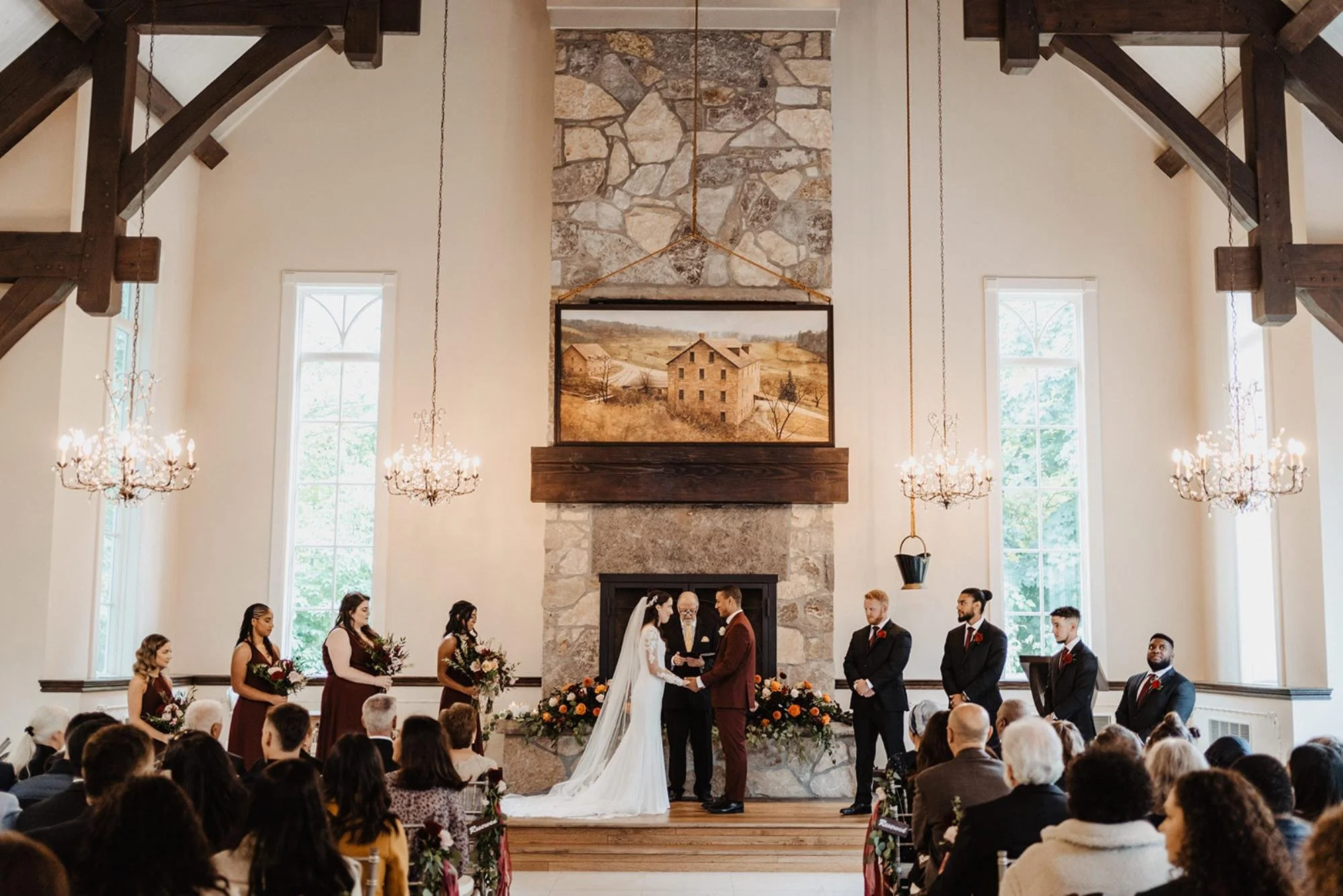 Bride and groom standing at the altar holding hands during their ceremony, surrounded by their wedding party in a bright chapel.