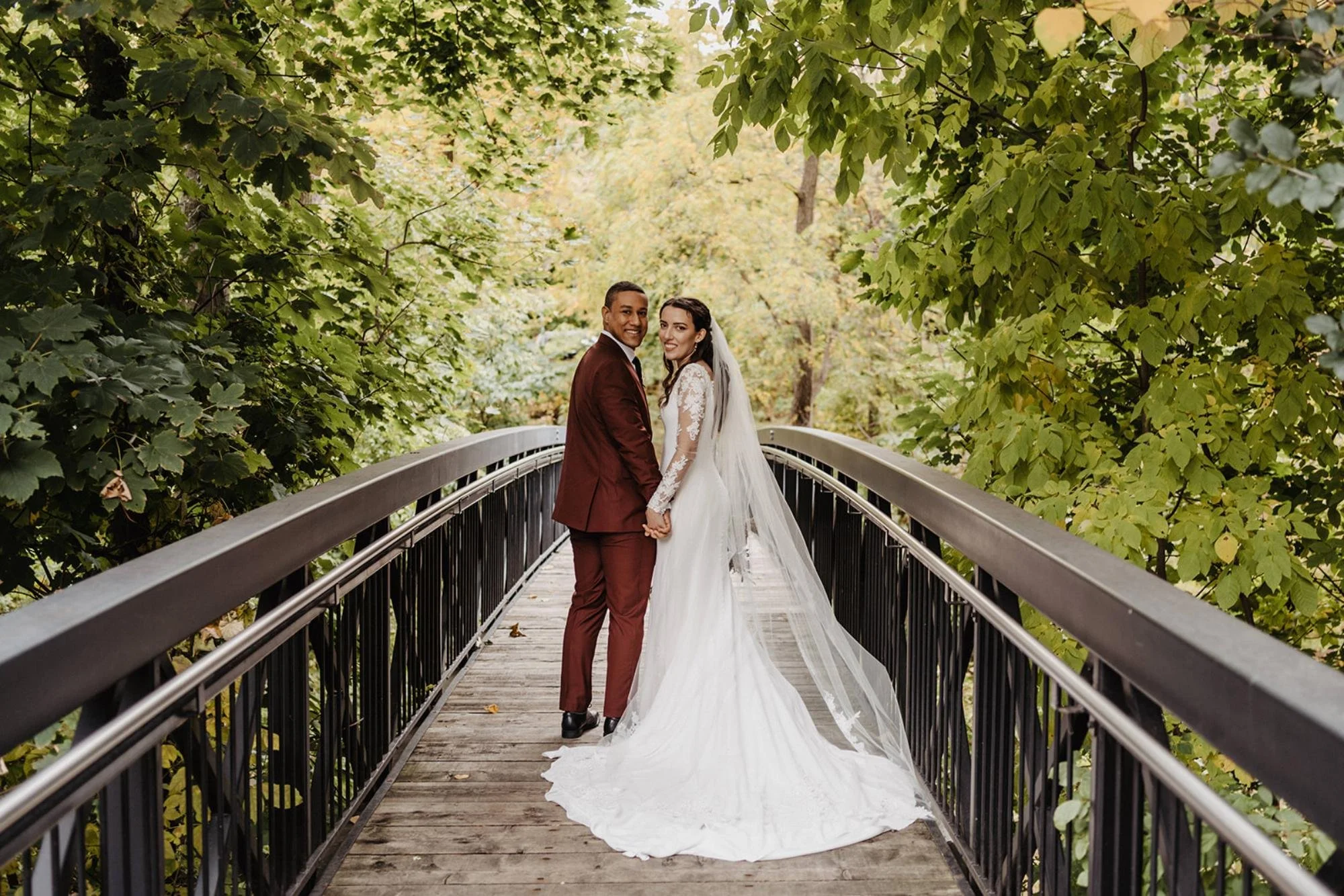 Bride and groom standing together on a wooden bridge surrounded by lush green trees.