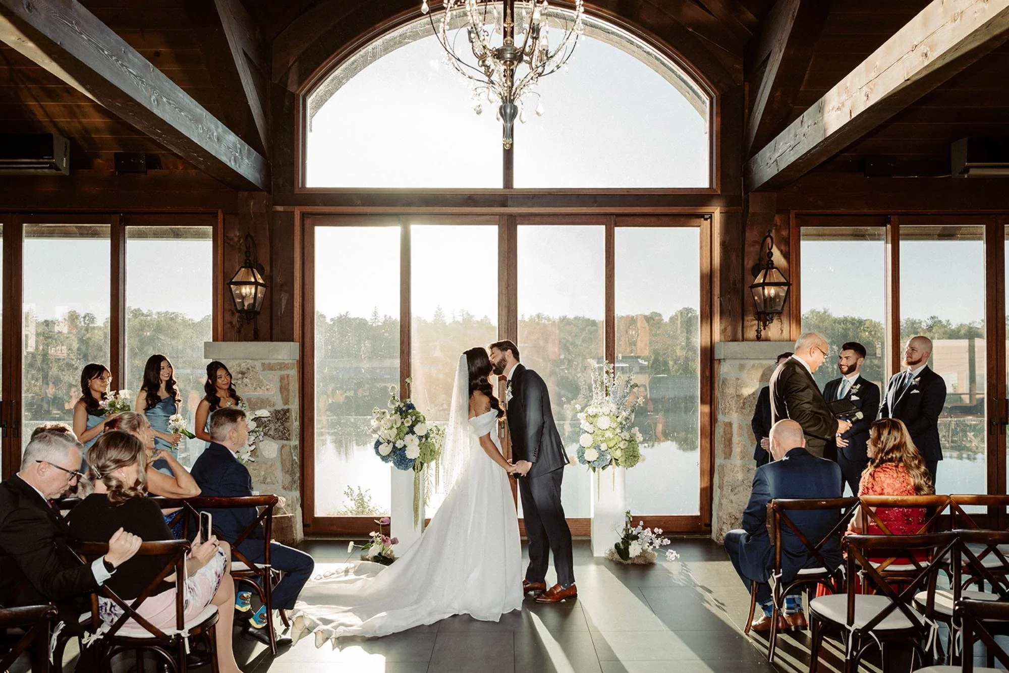 Bride and groom sharing their first kiss in front of floor-to-ceiling windows as guests watch in the sunlit ceremony space.