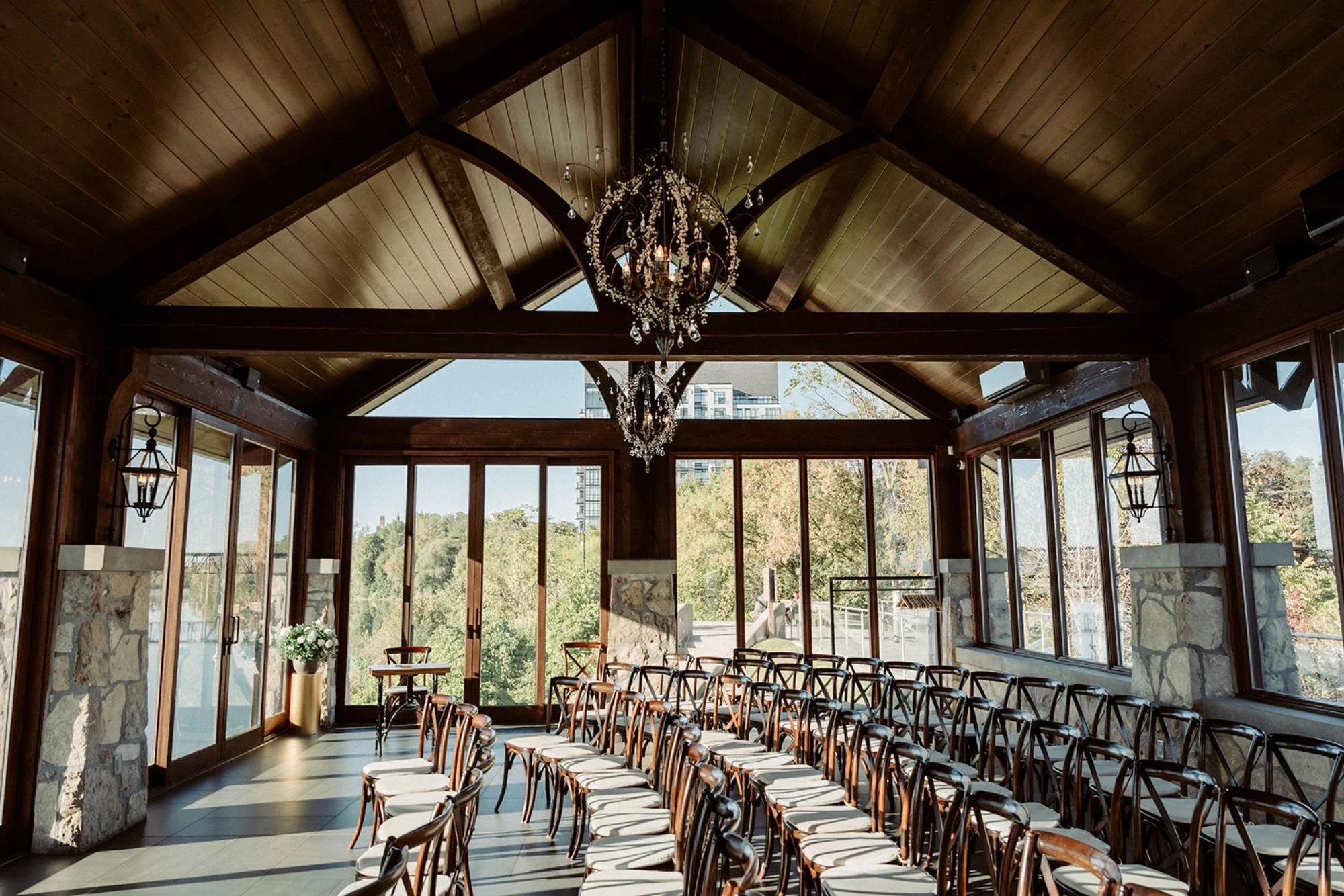 Indoor wedding ceremony space with wooden beams, large windows overlooking trees, and rows of wooden chairs facing the front.