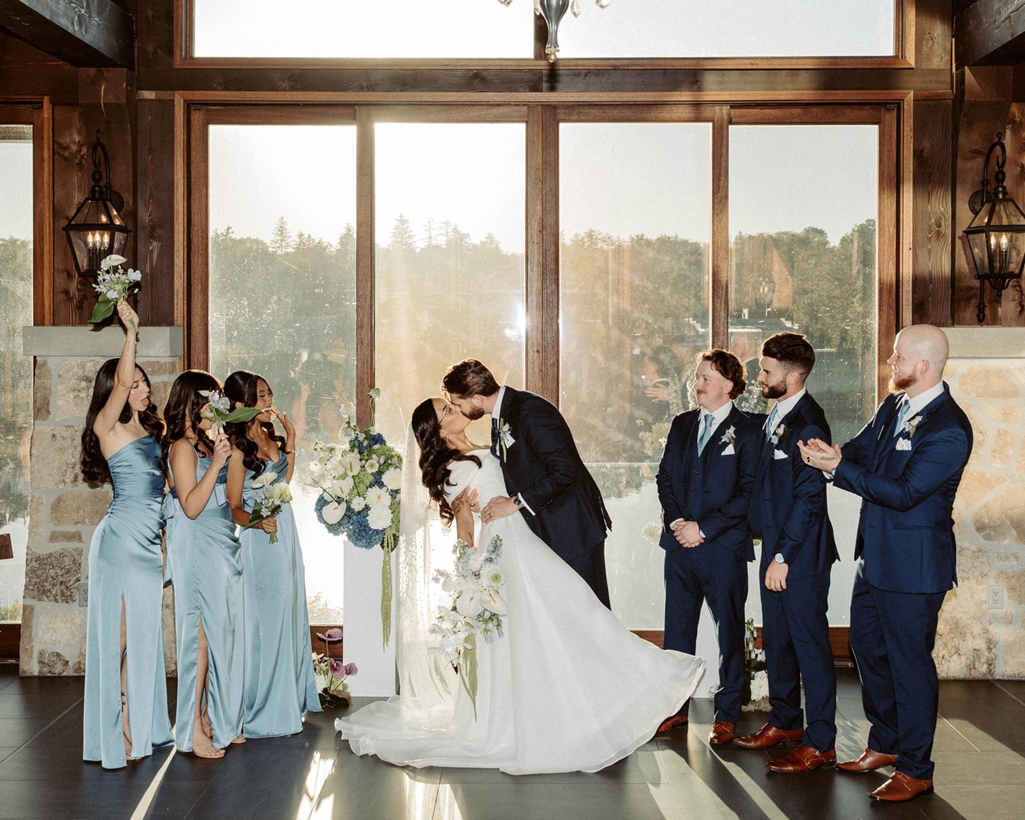 Bride and groom kissing at the altar while bridesmaids cheer and groomsmen applaud in a bright ceremony room.