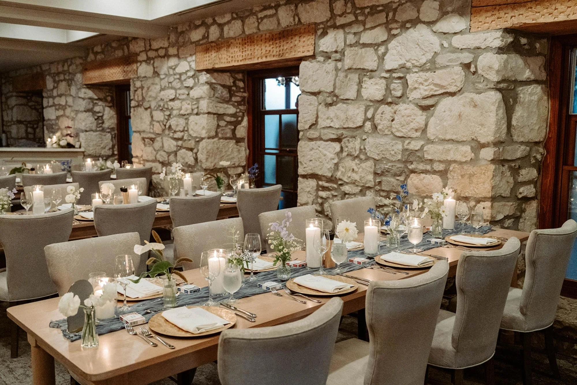 Reception room with stone walls and long dining tables decorated with candles, flowers, glassware, and place settings.