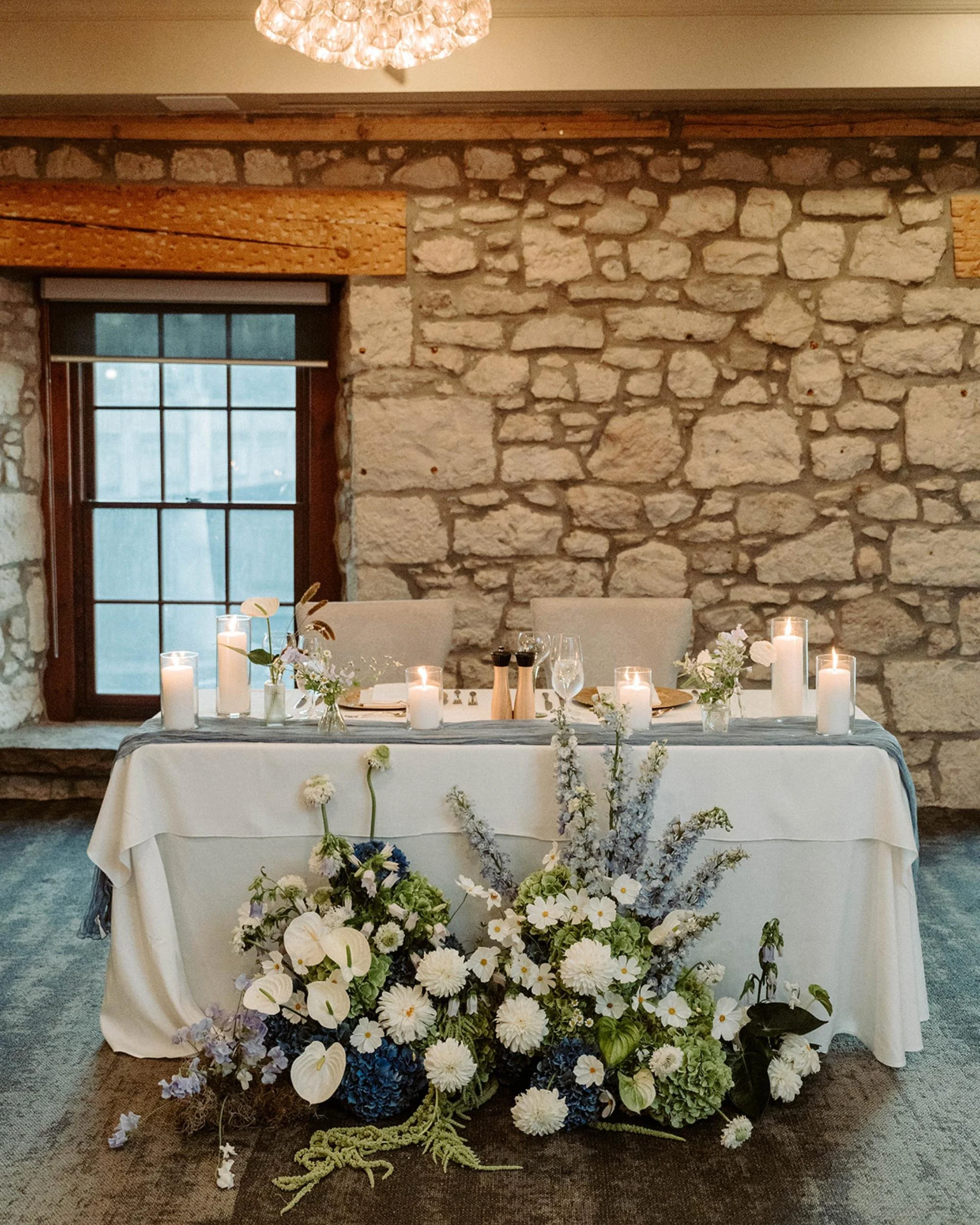 Sweetheart table set with candles and floral arrangements in front of a stone wall featuring blue and white blooms.