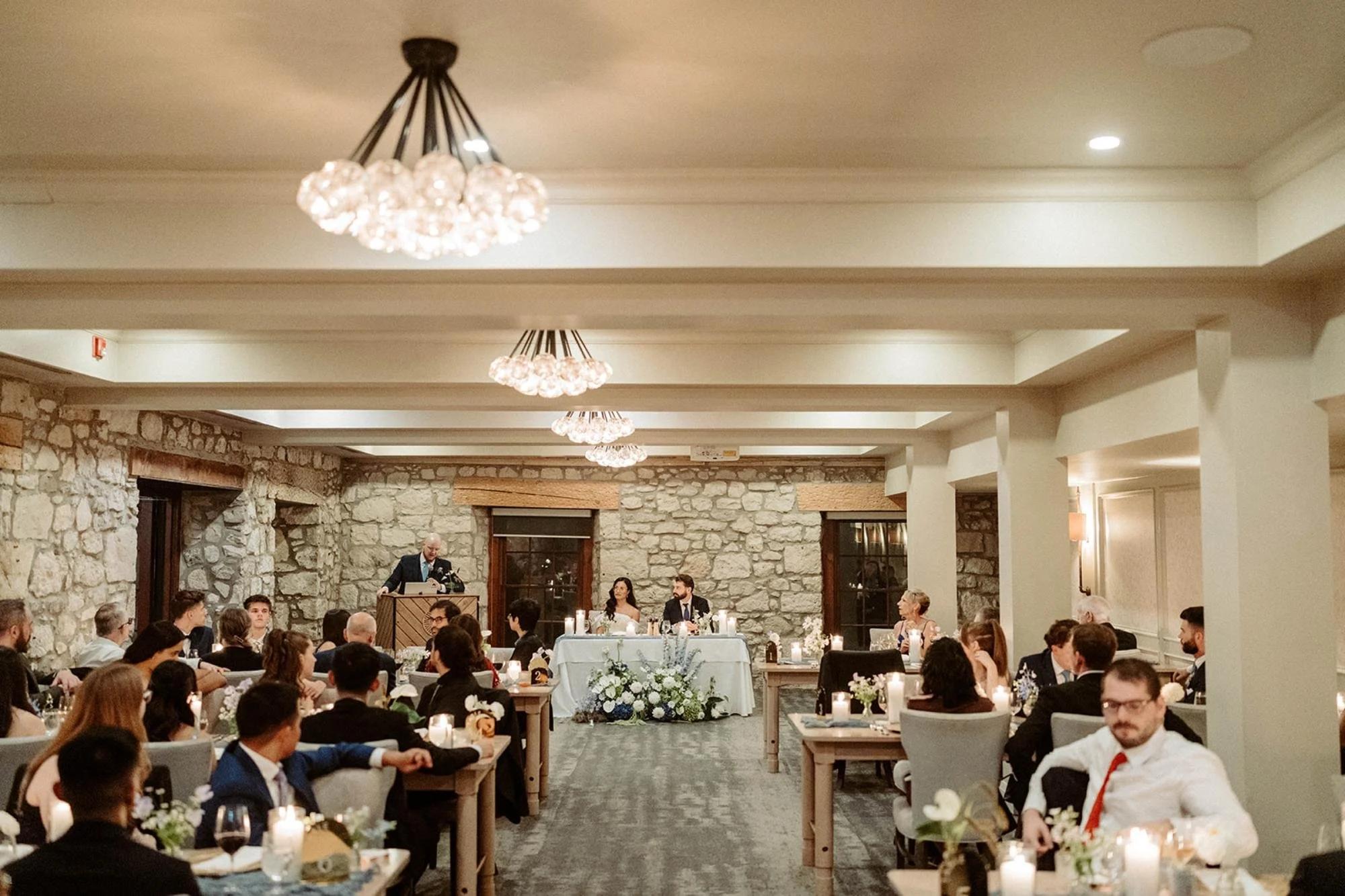 Wedding reception scene with a speaker at a podium and the bride and groom seated at the sweetheart table while guests watch.