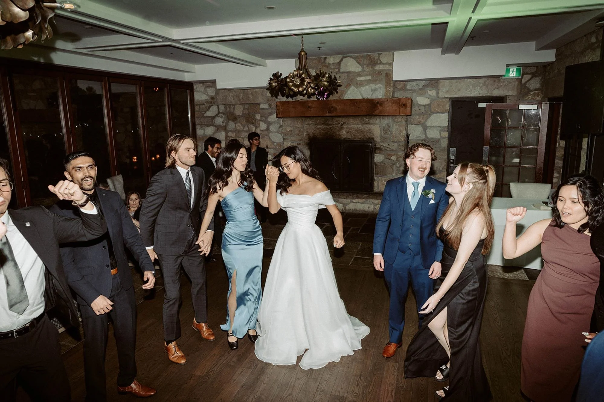 Bride, groom, and guests dancing together on the reception dance floor in a rustic room with stone walls.