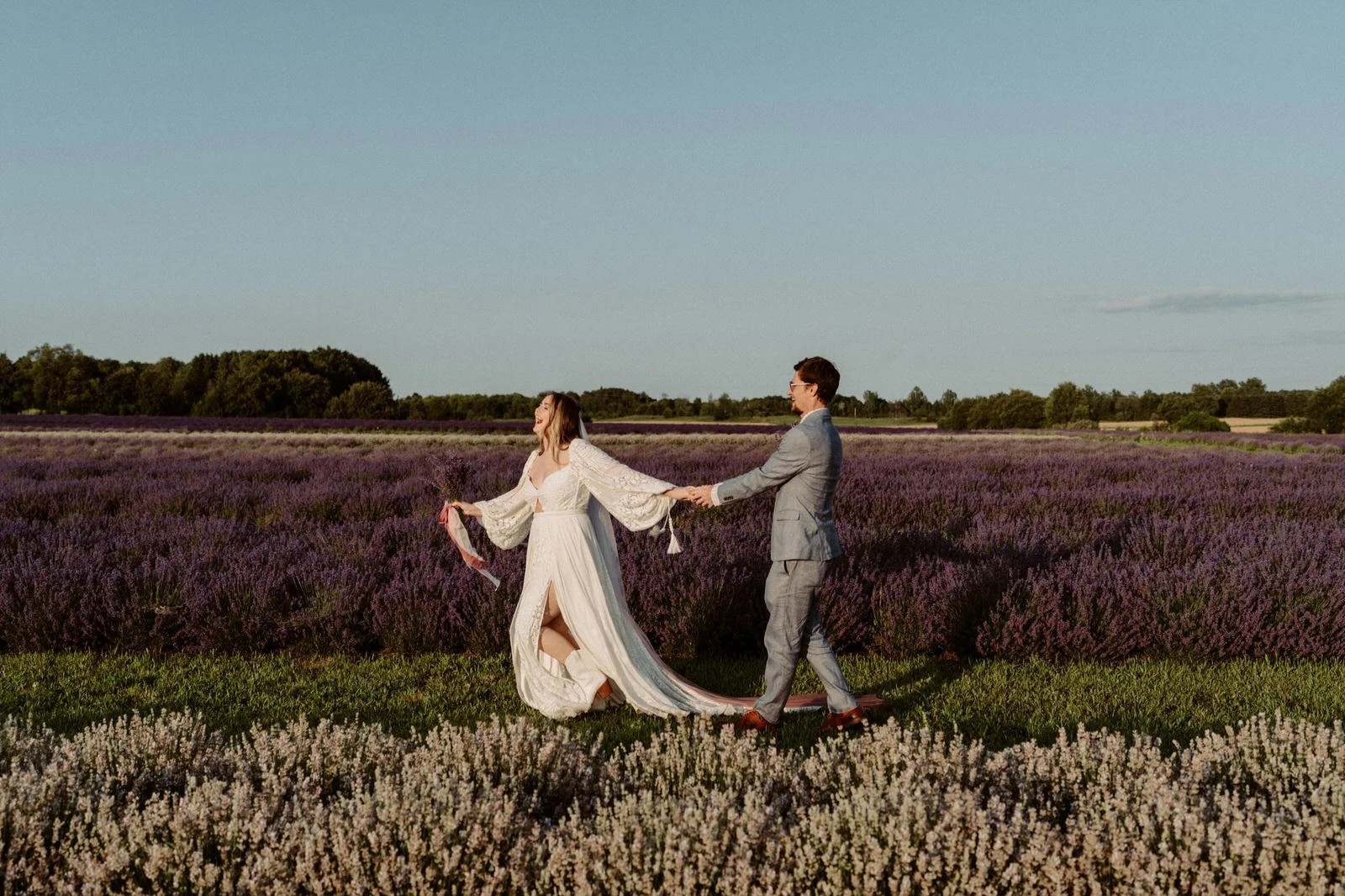 A couple in wedding attire holding hands and smiling in a lavender field with a clear blue sky.