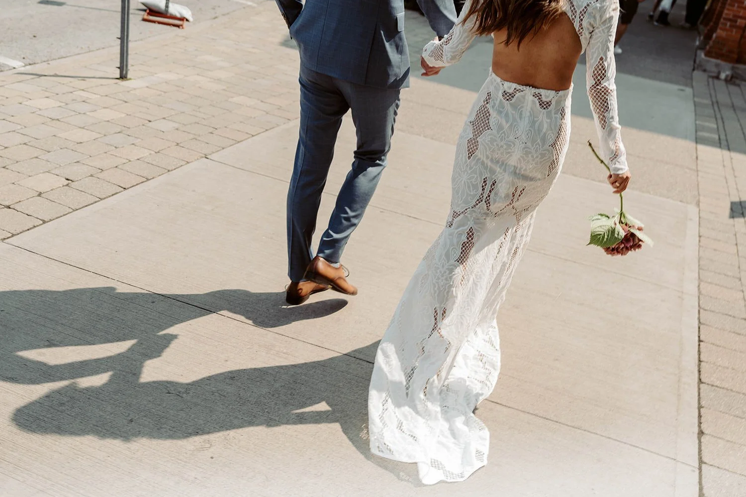 A couple walking on a sidewalk, the woman holding a flower and wearing a white lace dress, and the man wearing a gray suit.