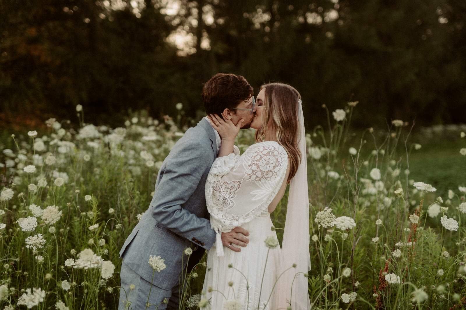 A couple kissing in a field of white flowers with trees in the background, during sunset.