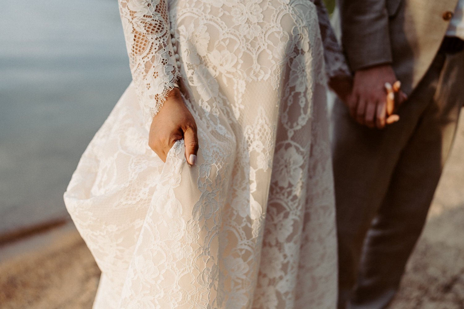 A bride and groom hold hands, with the bride wearing a lace wedding dress and the groom in a dark suit. The focus is on their hands and the details of their attire.