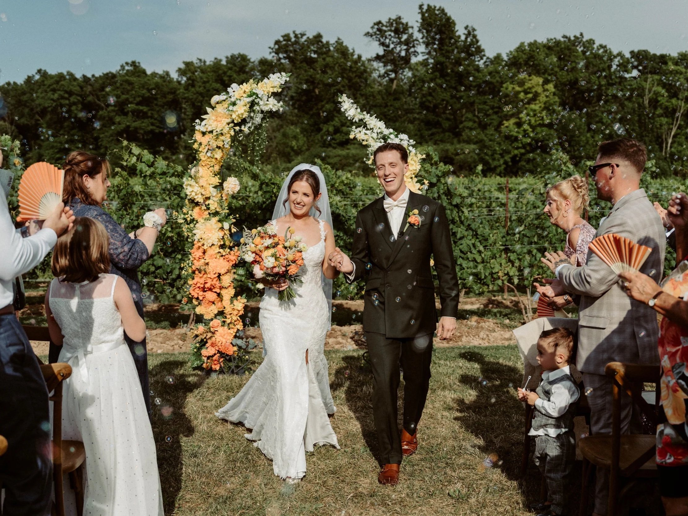 A bride and groom walking hand in hand through a group of wedding guests outdoors during daytime, with a floral arch behind them, celebrating their wedding.